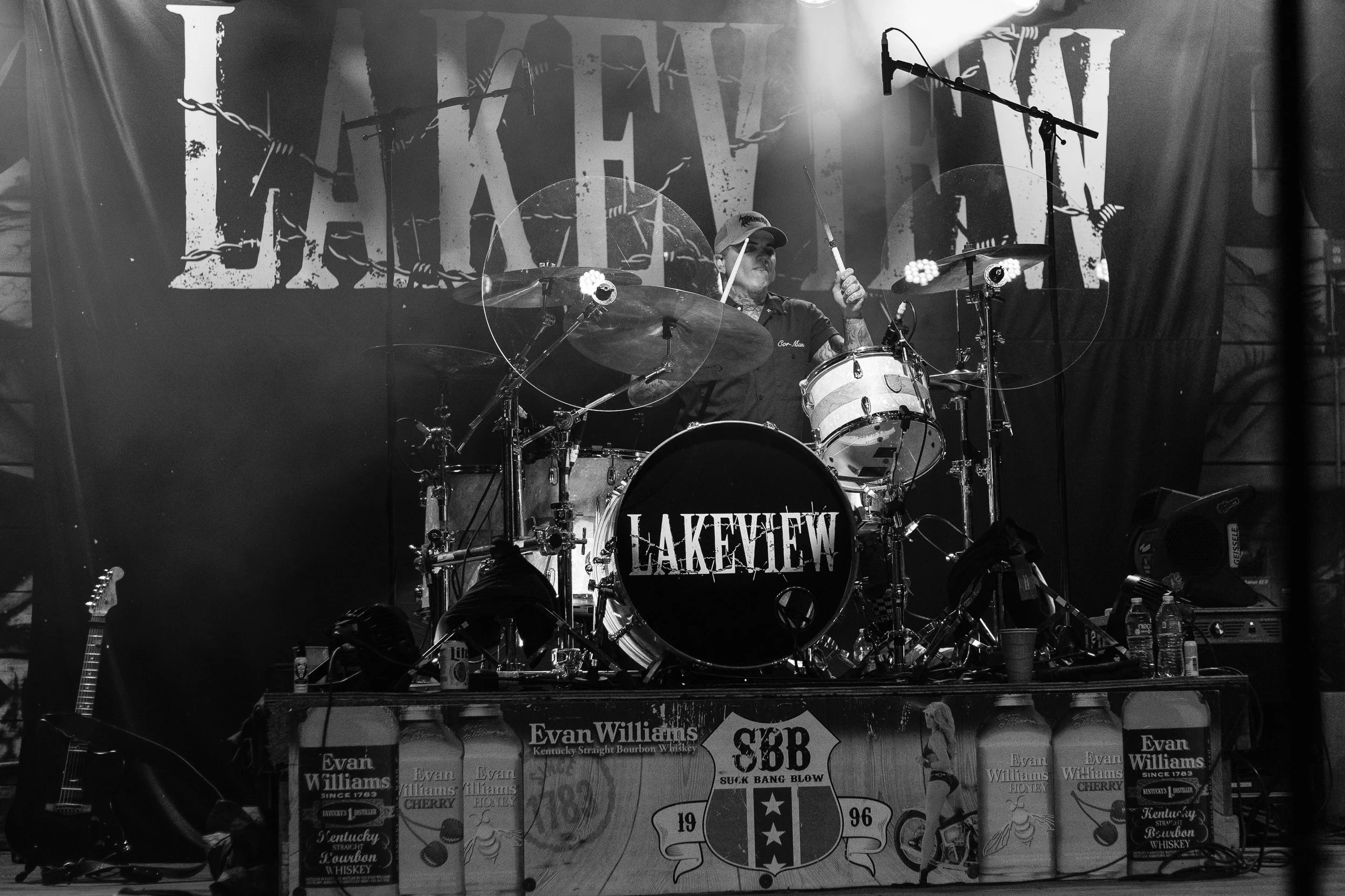 A musician playing drums on stage at a concert. The backdrop displays the word 'LAKEVIEW' in large, distressed letters. The stage is filled with various equipment and drinks.