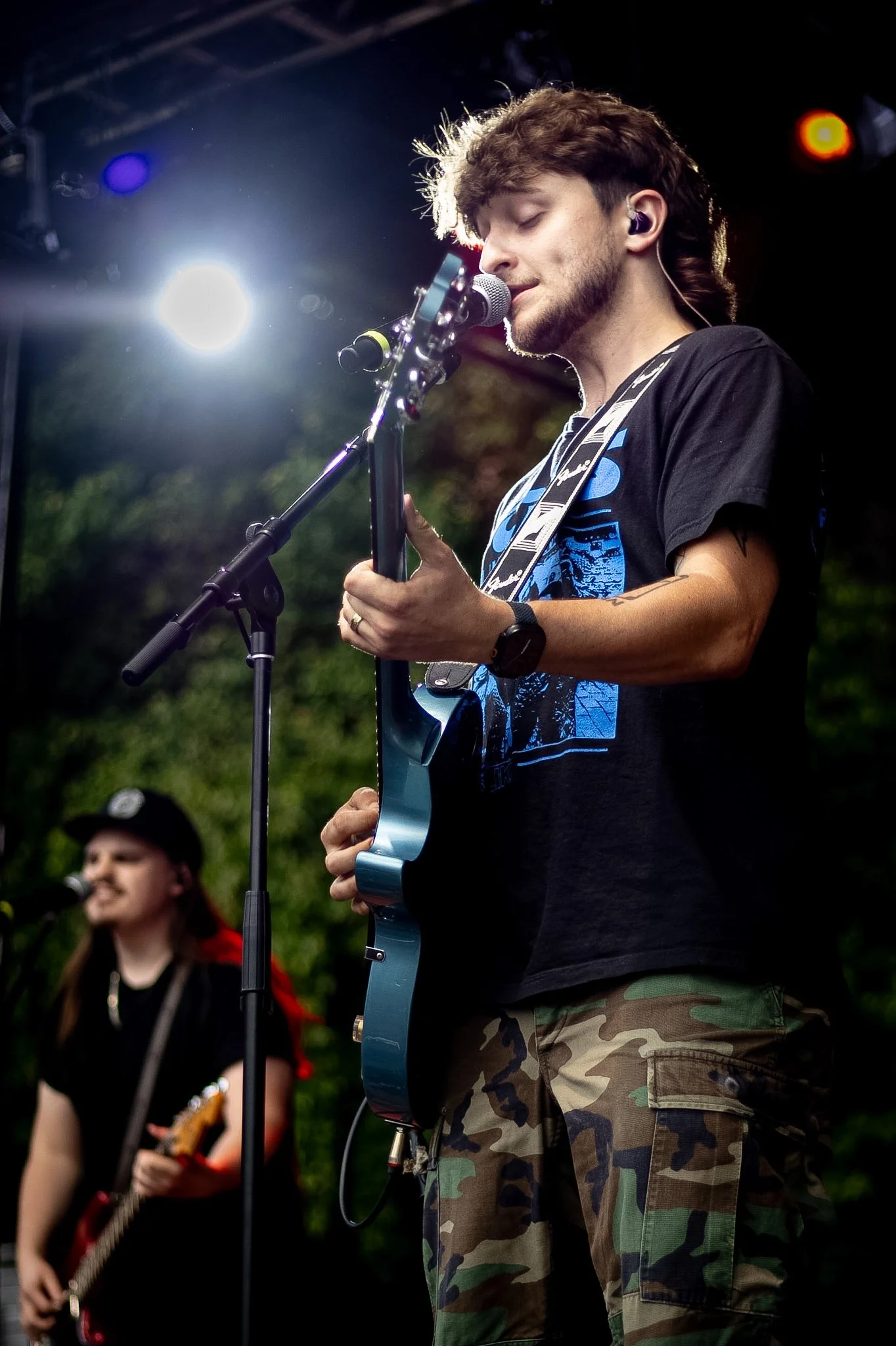 Two young musicians performing outdoors, one playing an electric guitar and singing into a microphone, with sunlight and stage equipment in the background.