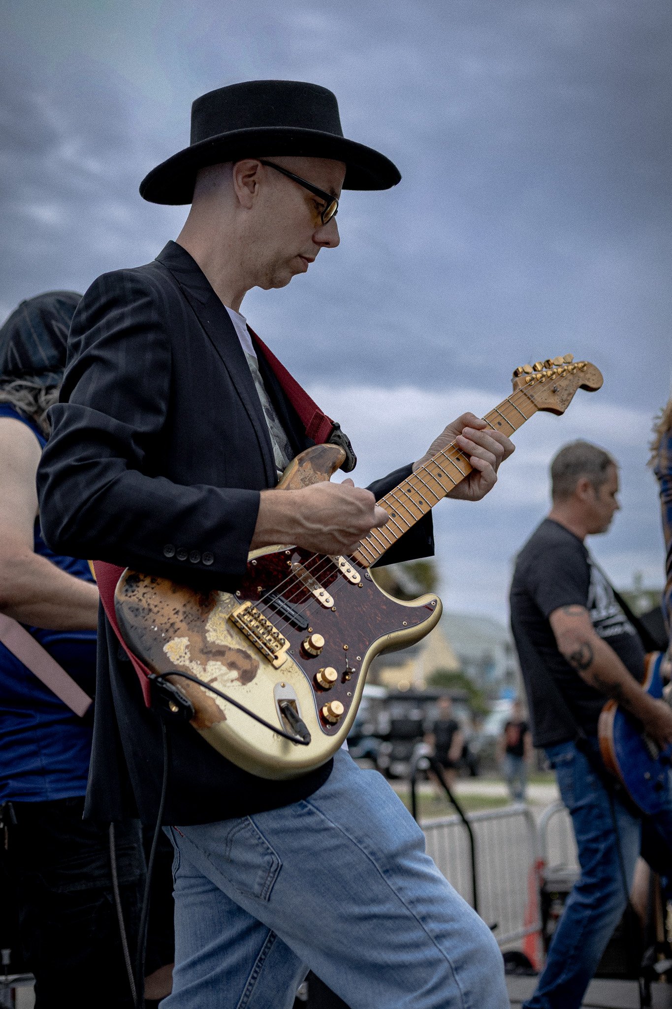 A man wearing glasses, a black hat, and a dark blazer is playing an electric guitar outdoors during daytime. Other people are visible in the background.