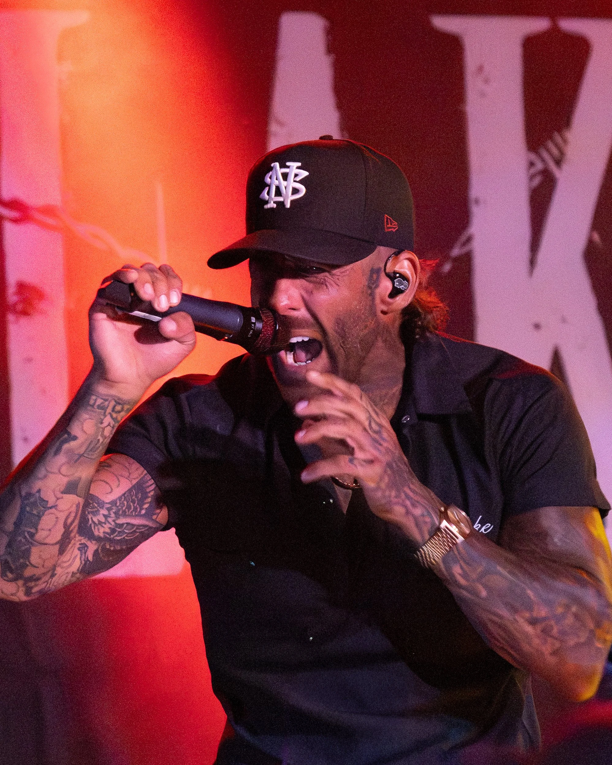 A man wearing a black cap with a white logo, singing passionately into a microphone, with tattoos on his arms, a gold watch, and an in-ear monitor.