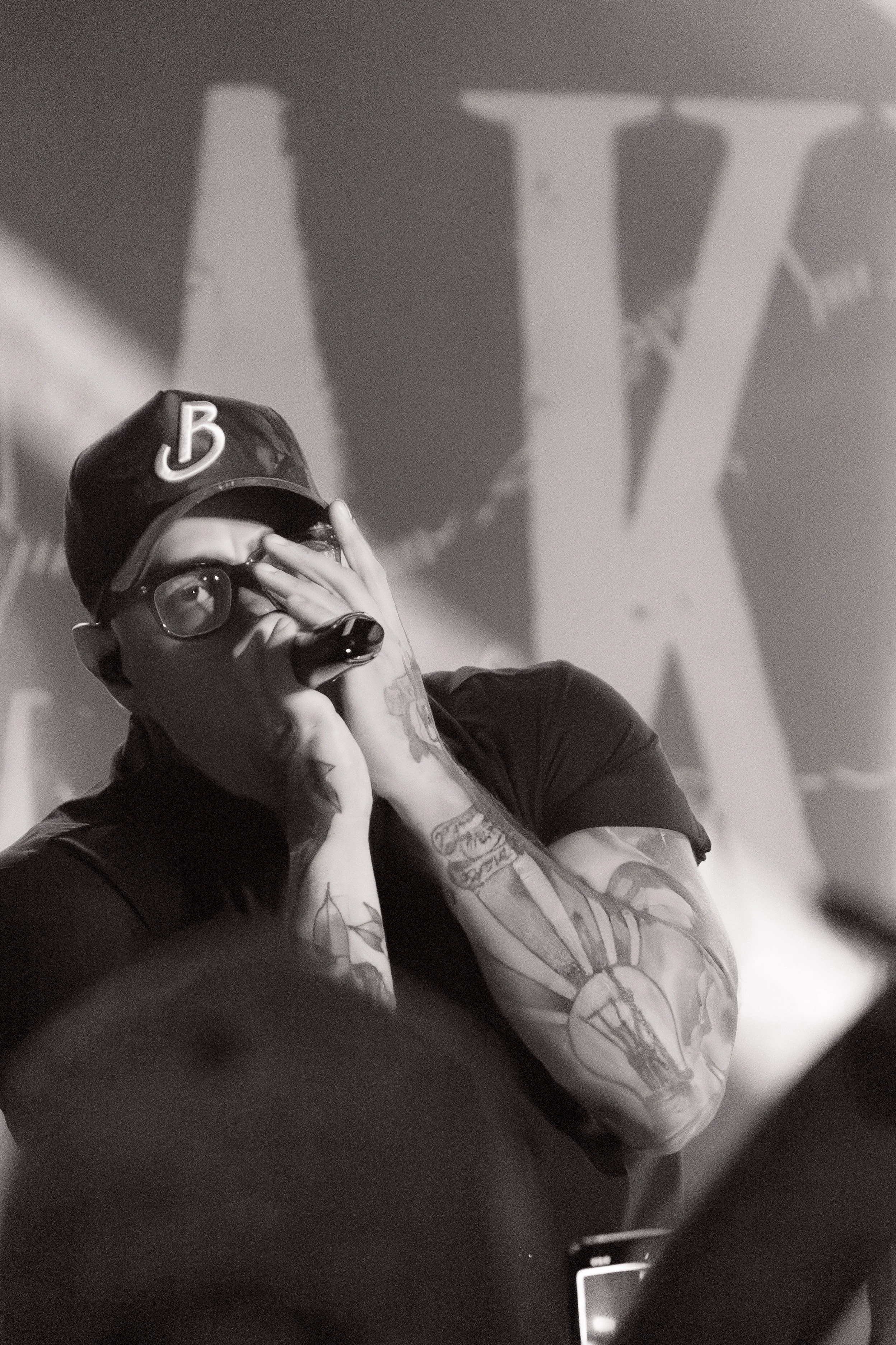 A person with tattoos on their arm and glasses, wearing a cap with a letter B and a Nike swoosh, is posing with their hand covering part of their face. The background features a large letter W and text.