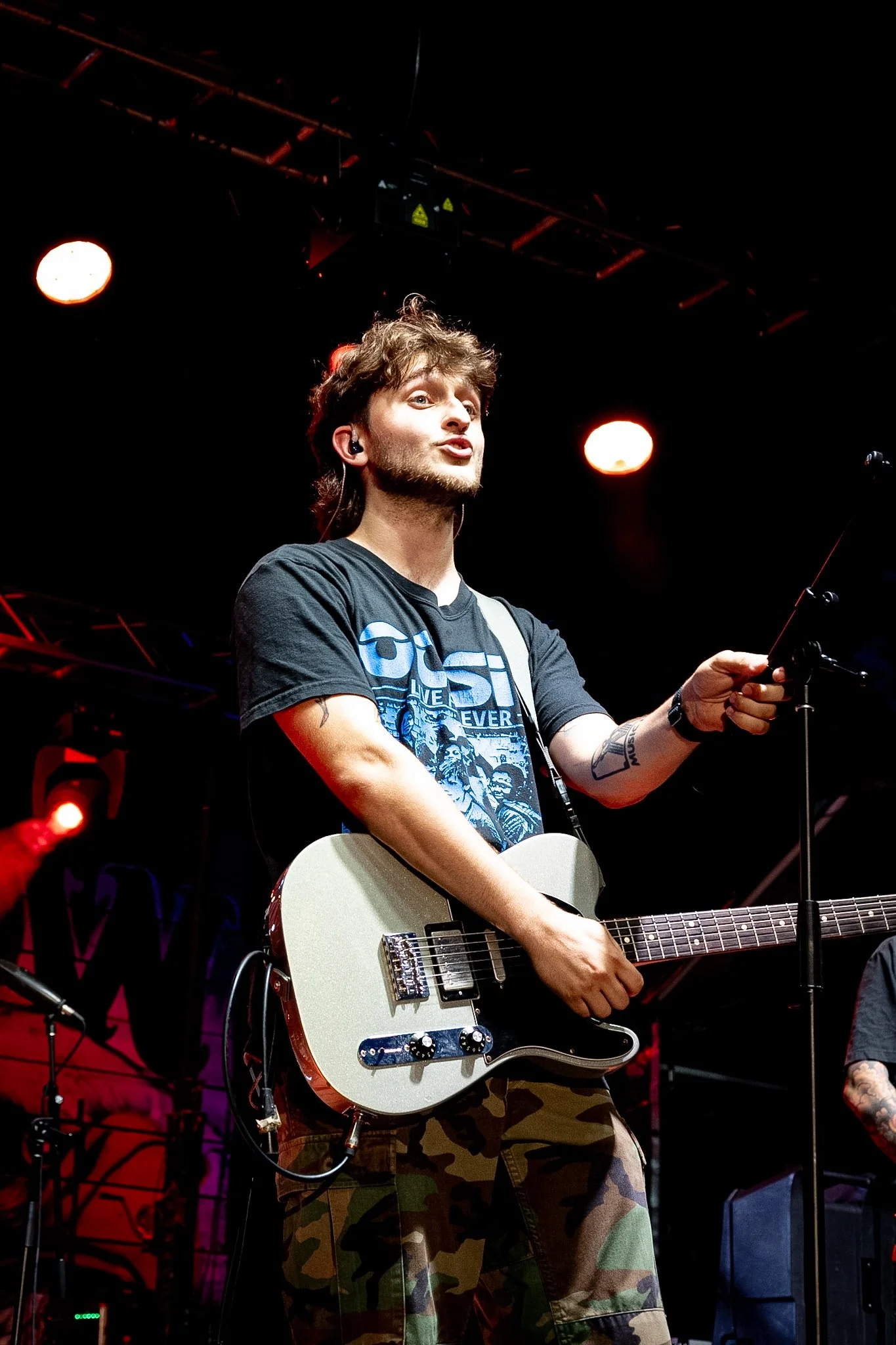 A young man with wavy brown hair and a beard playing an electric guitar on stage under red and white stage lights.