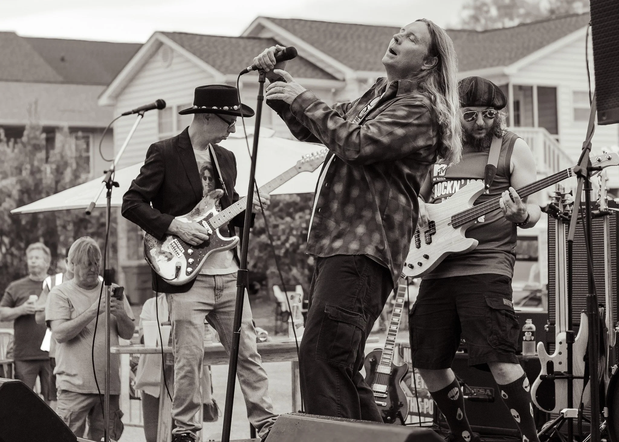 Black and white photo of a band performing outdoors; a woman singing into a microphone, two guitarists playing electric guitars, and an audience in the background.