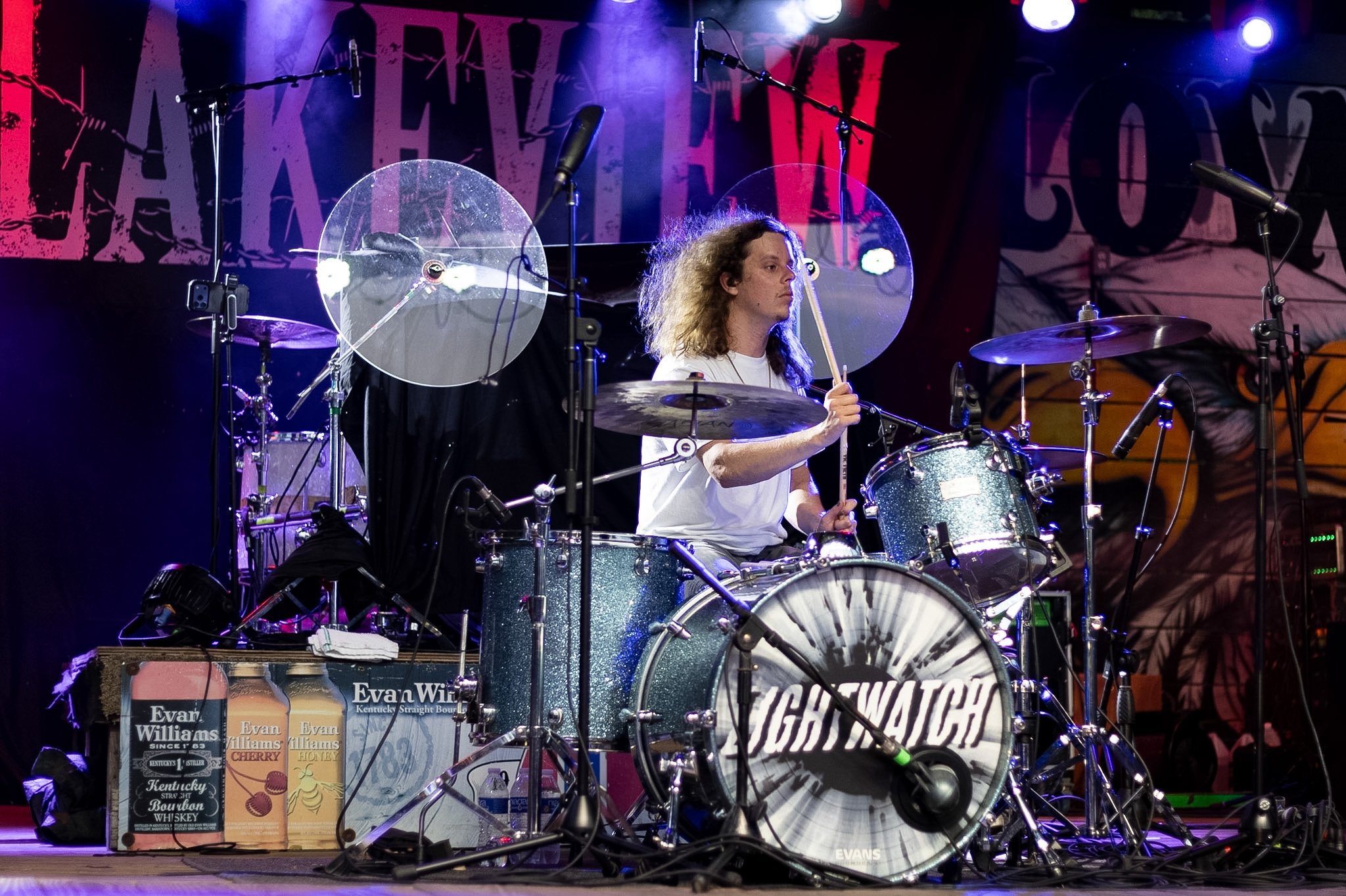 A drummer with long curly hair playing a drum kit on stage with microphones and lights, with posters and a banner in the background.