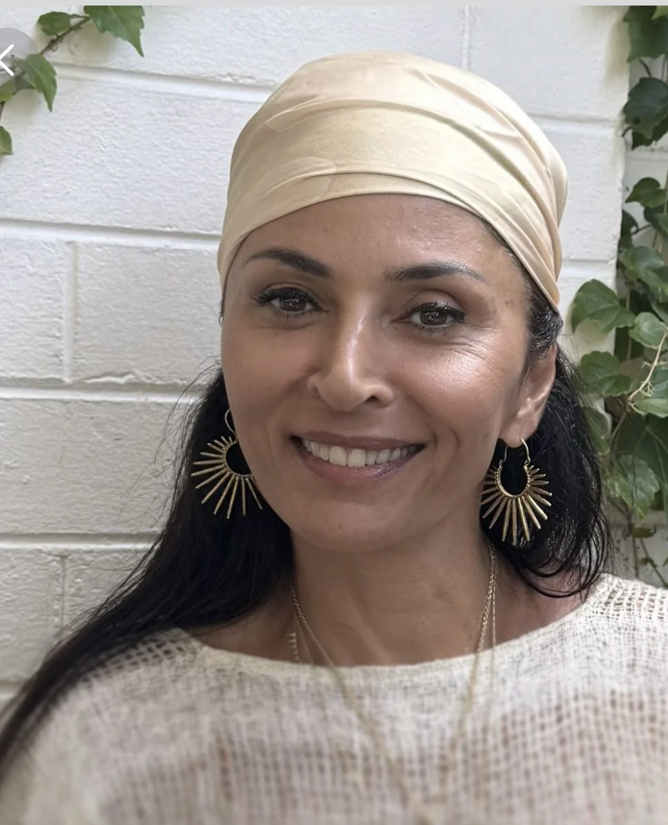 A smiling woman with long dark hair wearing a beige head wrap, gold dangling earrings, and a light-colored top, standing in front of a white brick wall with green foliage.