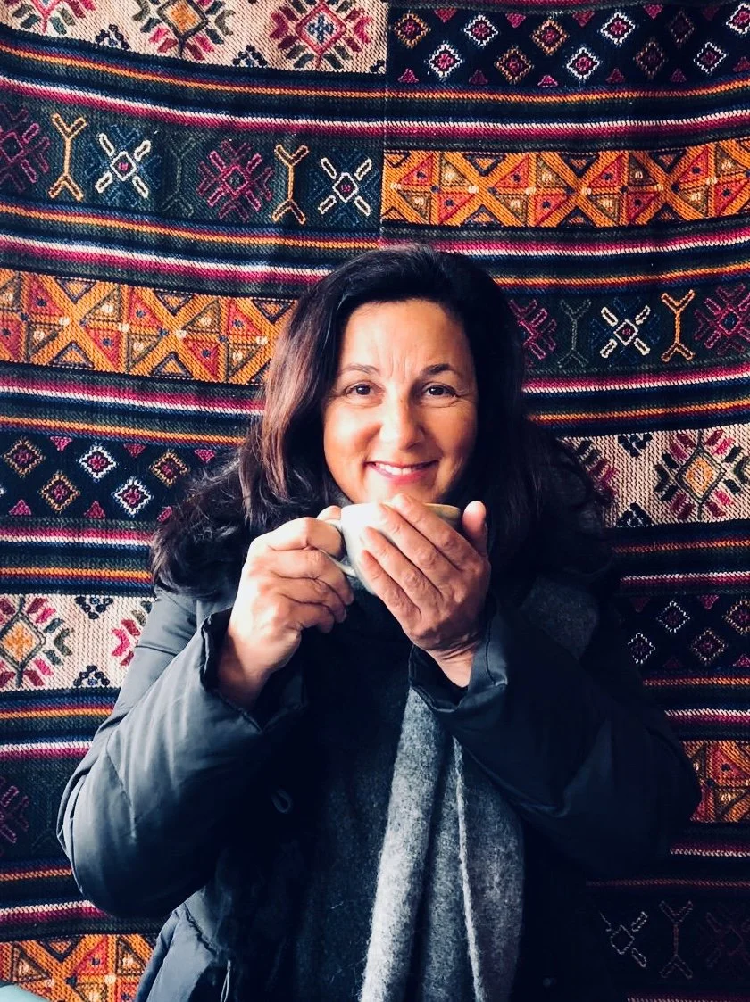 A woman smiling and holding a small cup, standing in front of a colorful, patterned textile background.