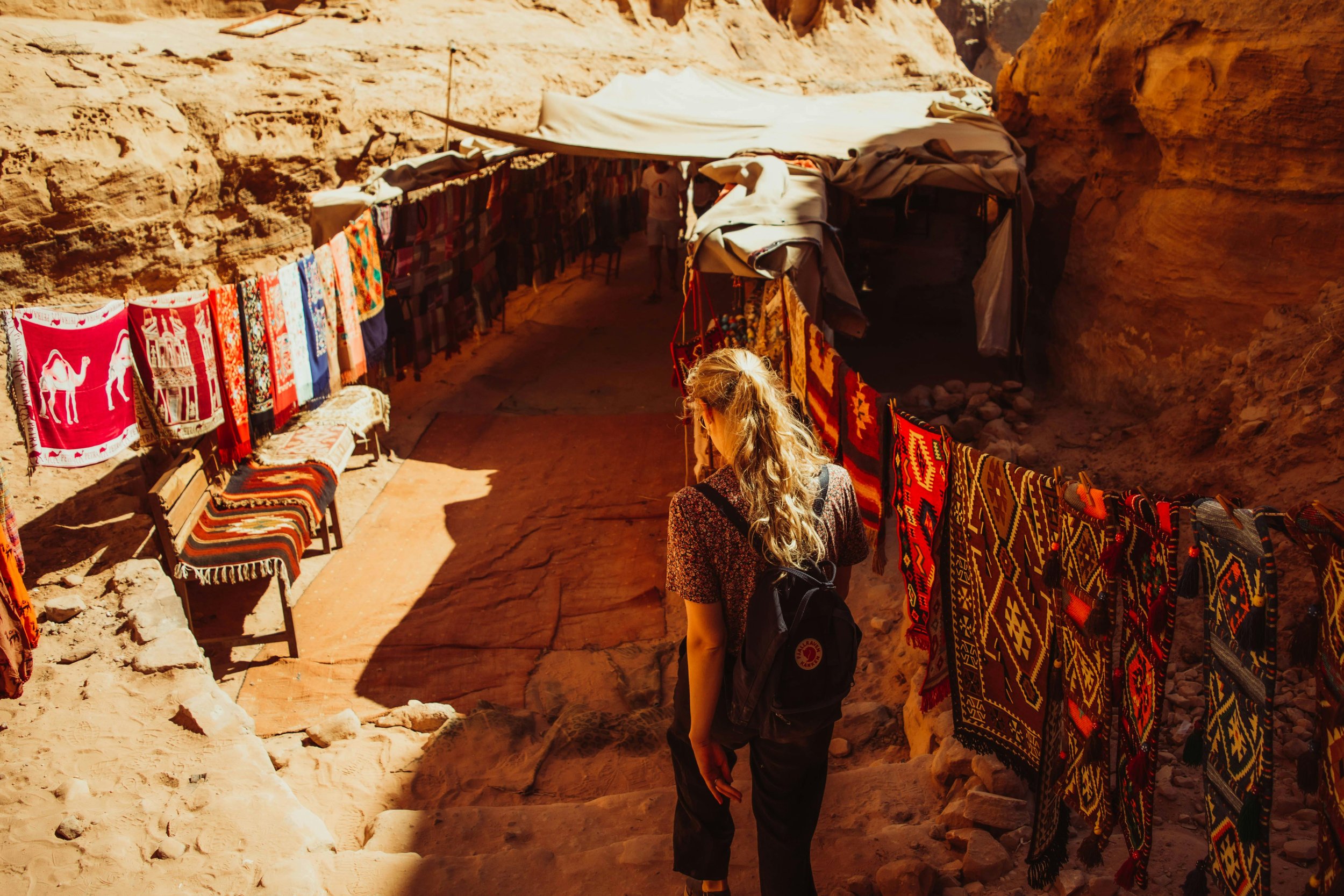 A woman with a backpack walking through a marketplace with colorful woven textiles hanging on both sides in a desert-like environment.