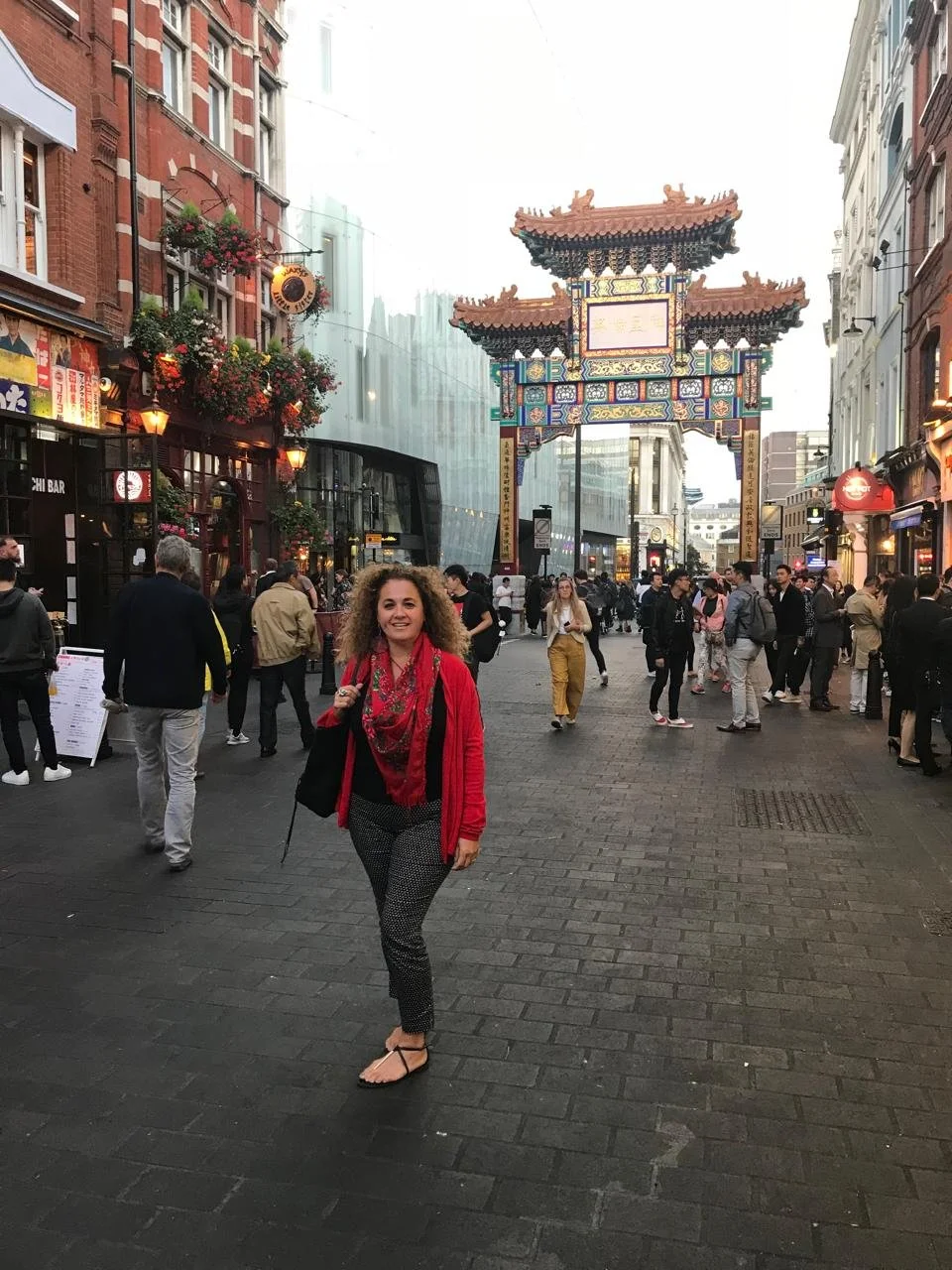 A woman with curly blonde hair, wearing a red jacket, black and white patterned pants, and flip flops, smiling while standing on a busy Chinatown street with a decorative Chinese archway overhead. People walk around in the background, with shops and buildings lining the street.