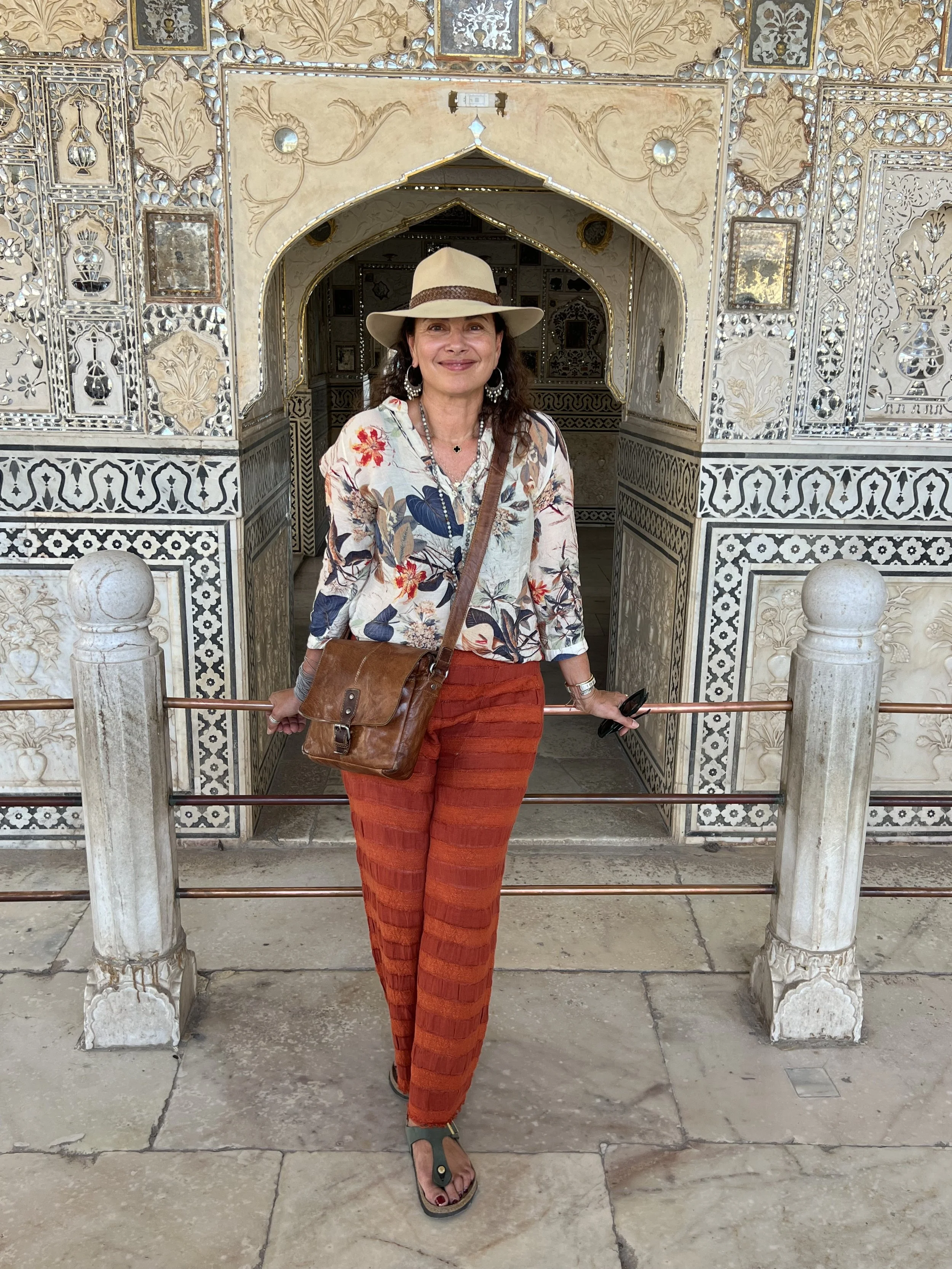 Woman standing in front of ornate architecture with intricate carvings and patterns, wearing a beige hat, floral shirt, orange striped pants, and carrying a brown shoulder bag and sunglasses.