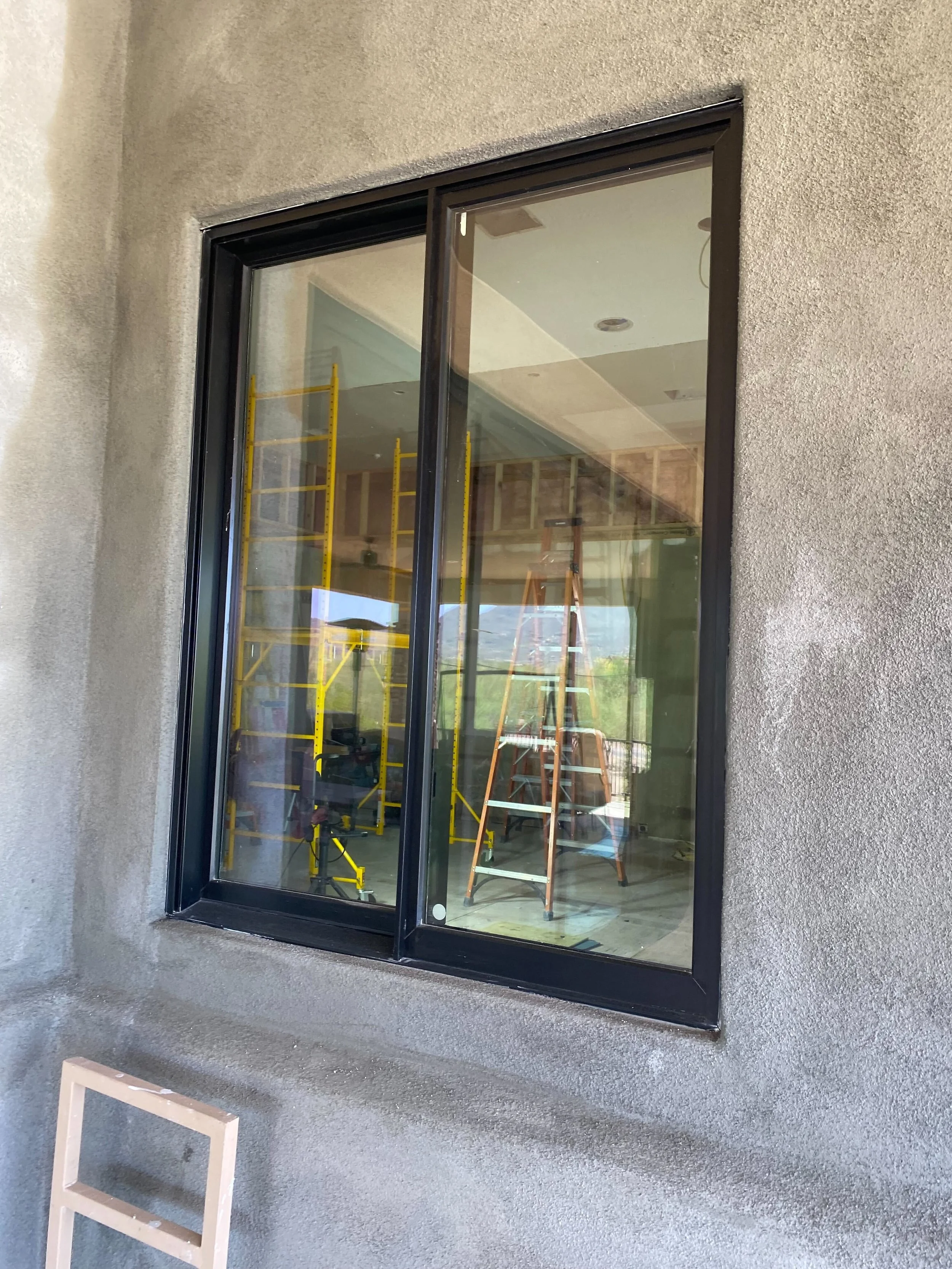 View of a construction site through a large exterior window showing ladders, scaffolding, and construction tools inside a partially finished room.