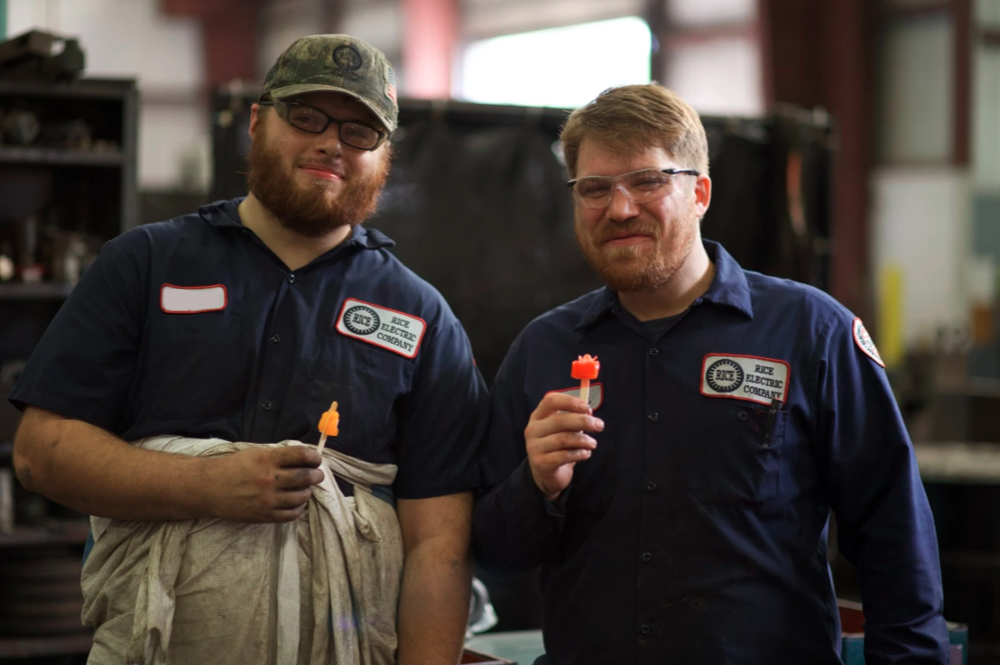 Two men in work uniforms holding small red and orange plastic objects, standing inside a workshop or industrial setting.