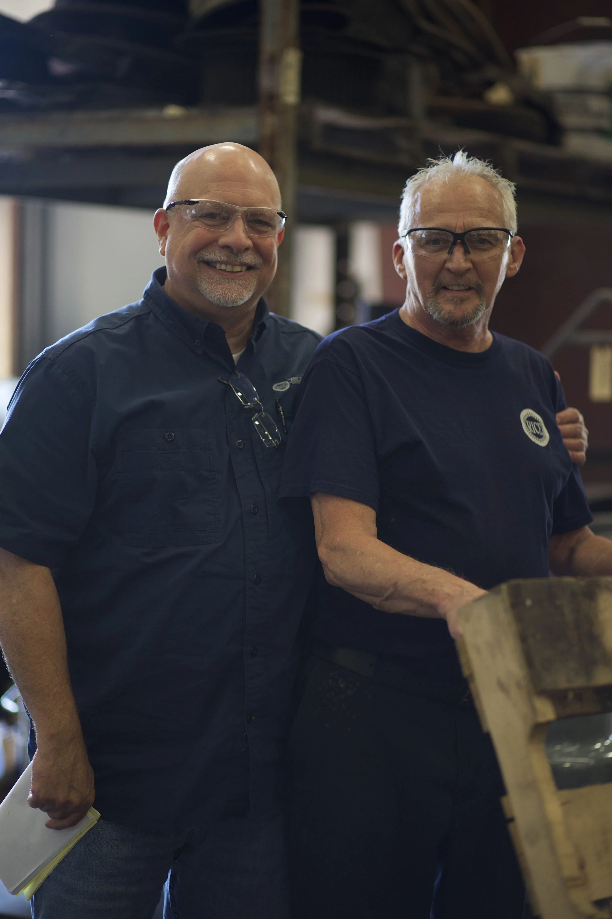 Two men smiling, wearing safety glasses in an industrial or workshop setting with metal shelves and equipment in the background.