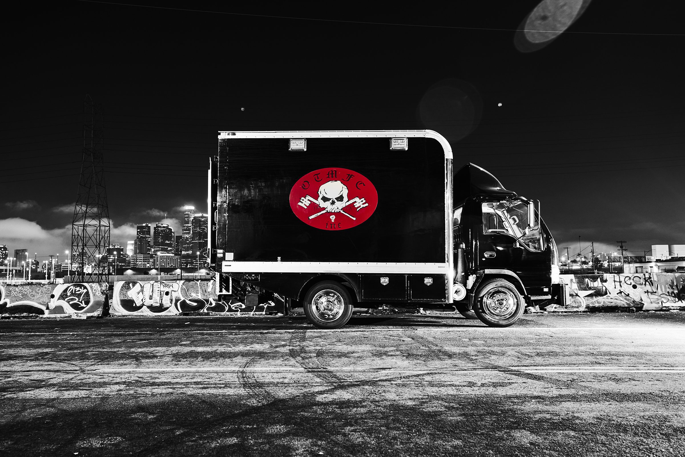 Black and white photo of a truck with a skull and crossbones logo parked near graffiti-covered barriers, city skyline and moon in background.