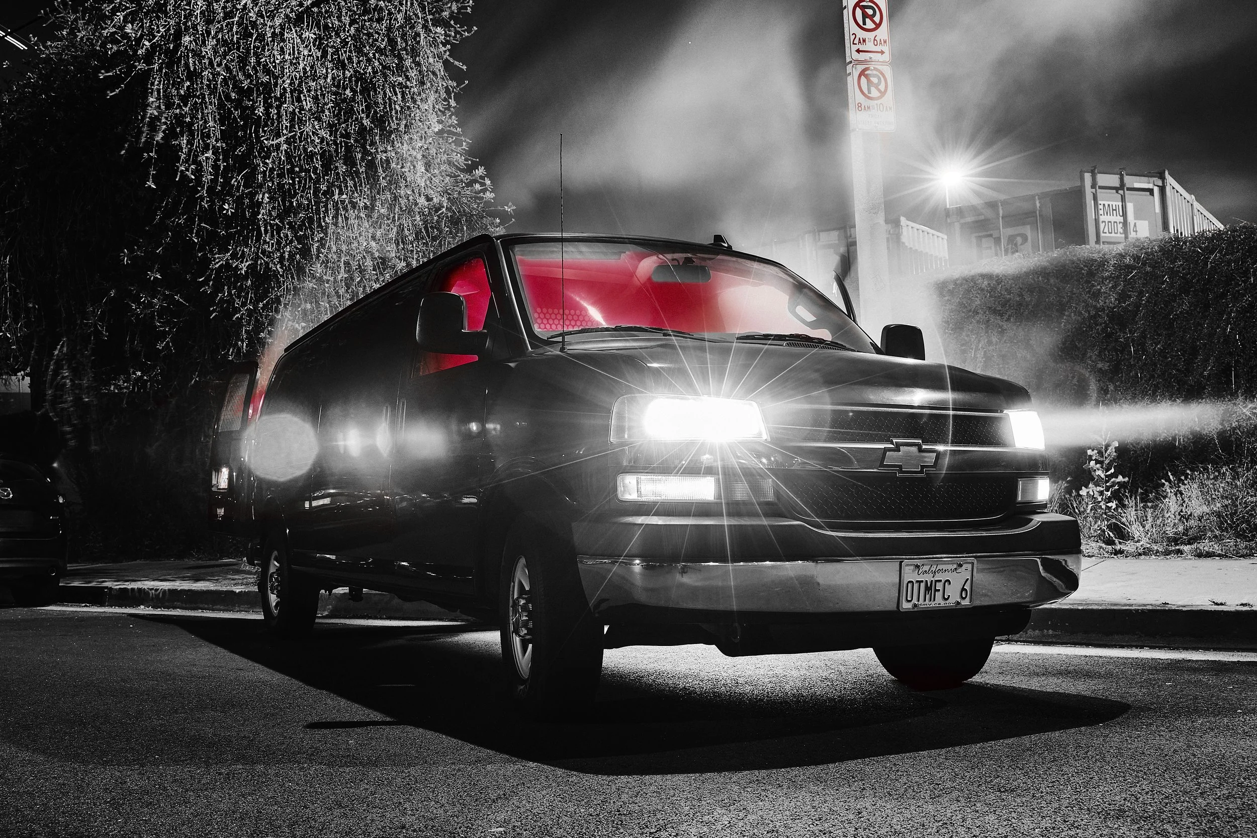 Black Chevrolet truck with bright headlights parked in a lot at night, behind a hedge, with a no parking sign and some containers in the background, illuminated by streetlights.