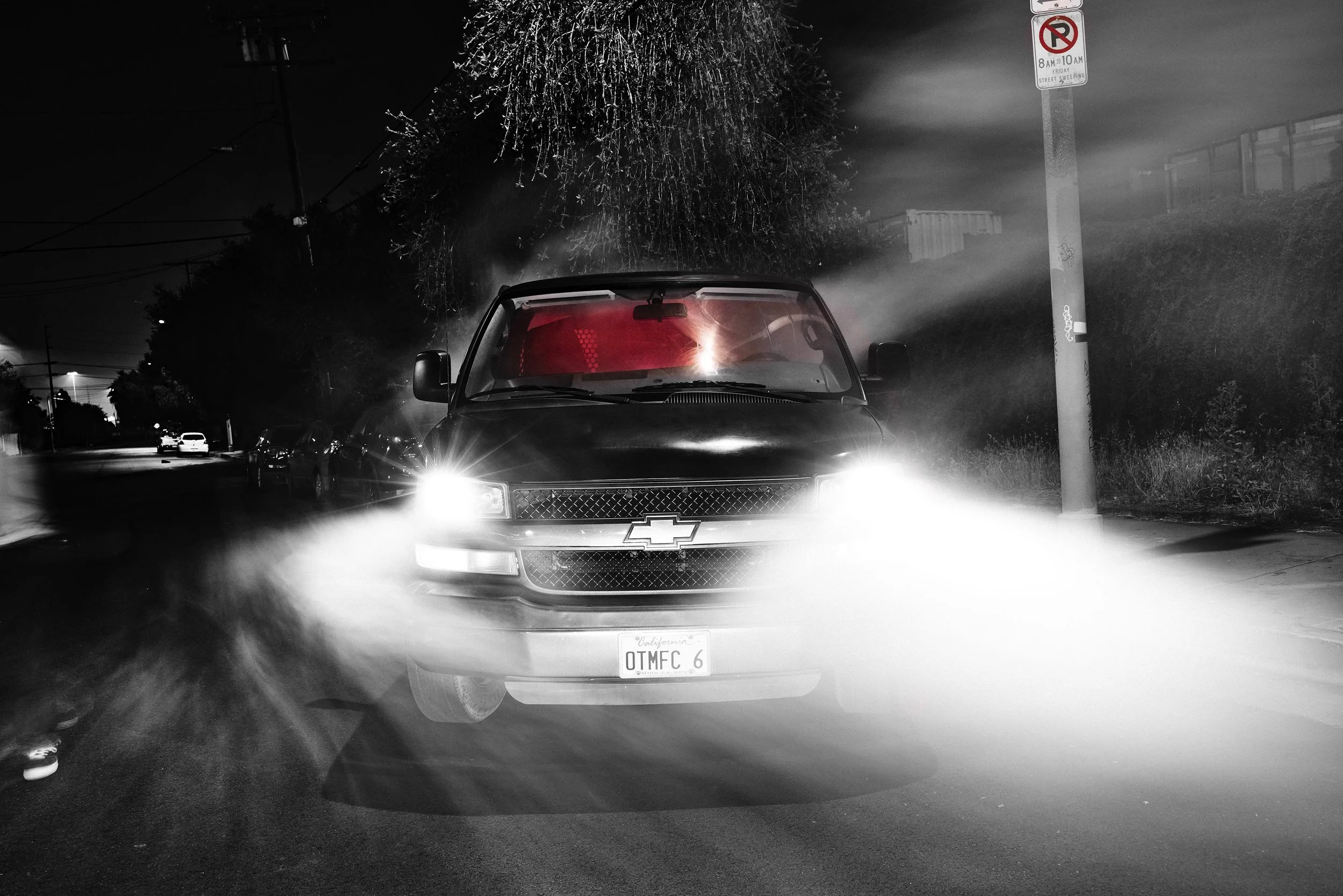 A black Chevrolet pickup truck with California license plate OTMFC6 driving at night. The headlights are on, and the street is misty, with a tree and a no parking sign on one side.