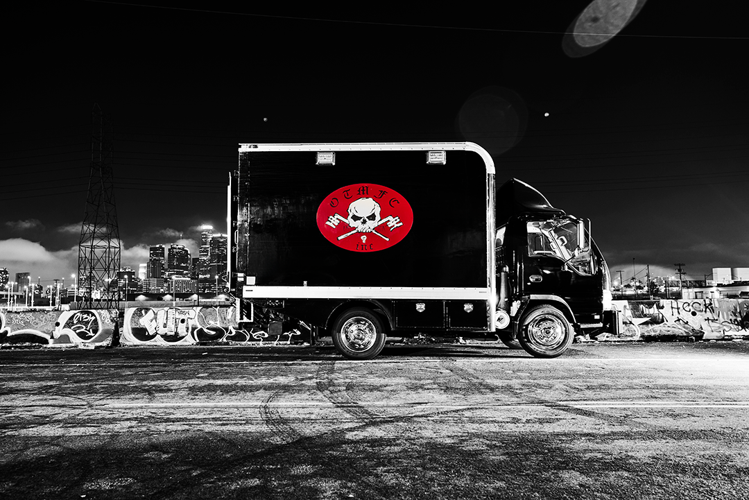 A black delivery truck with a red logo featuring a skull and crossed bones parked on a graffiti-covered street at night in an urban area with city buildings in the background.