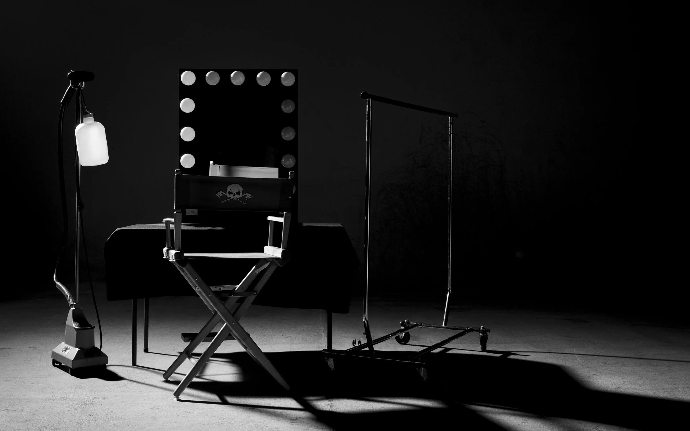 A black and white photograph of a makeup or dressing room setup with a director's chair featuring a skull and crossbones design, a mirror with round lights, a table with a bottle, a standing lamp, and a clothing rack against a dark background.