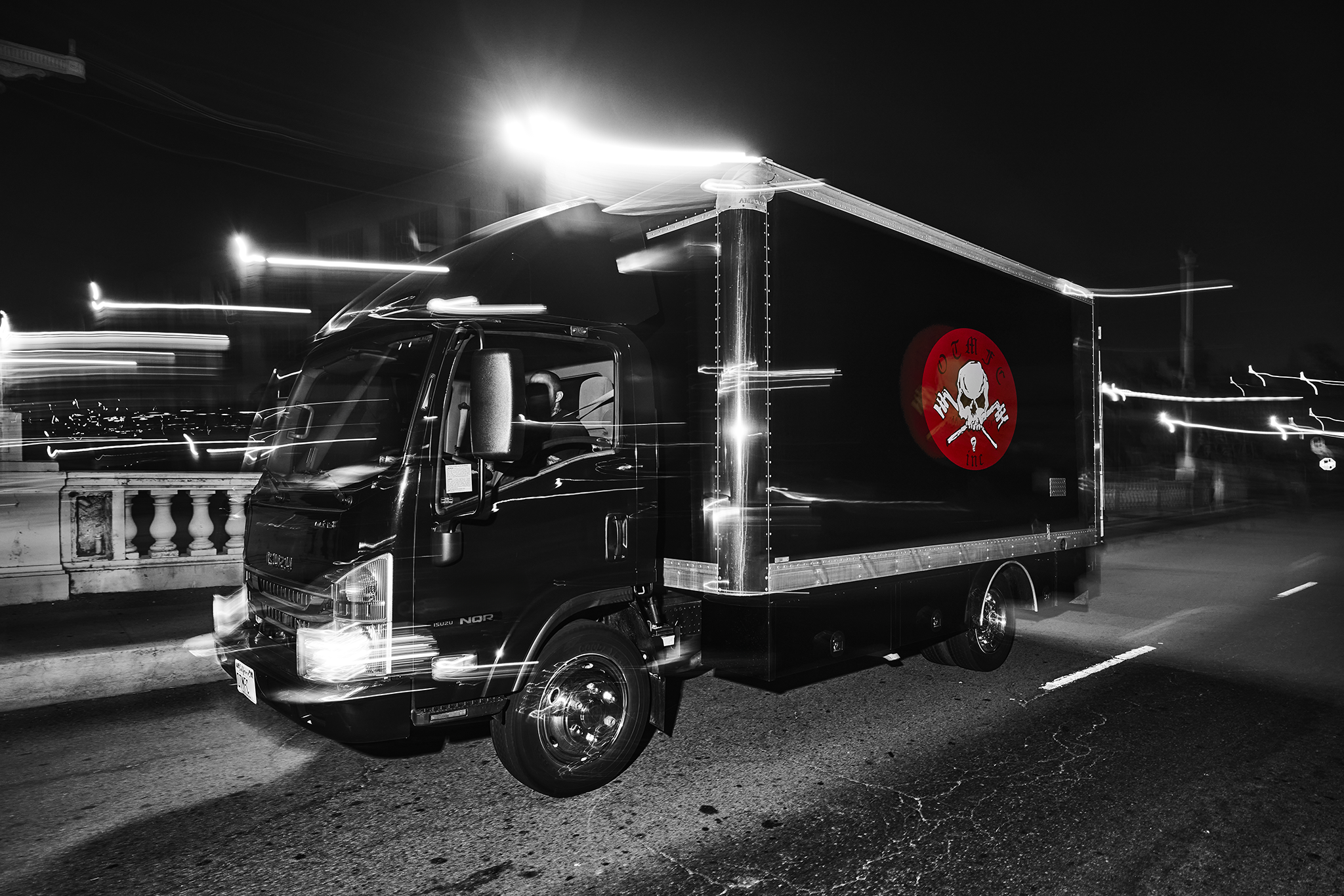 A black truck with a skull and crossbones logo on its side, parked in an urban area at night with city buildings and graffiti in the background.