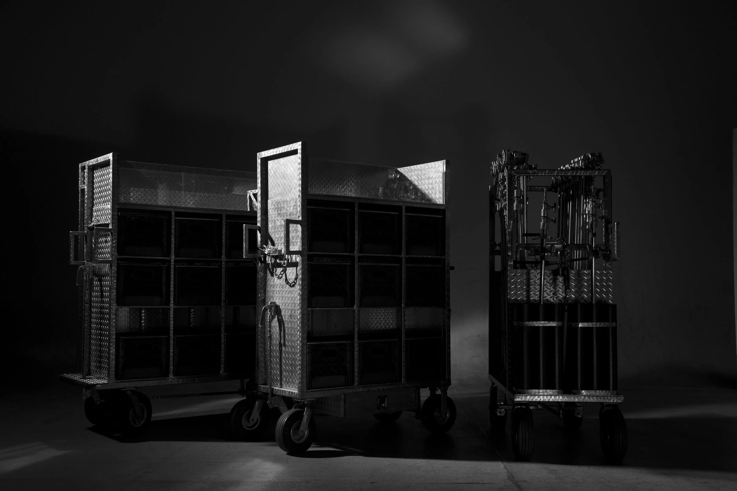 Black and white photo of various metal carts with open storage compartments, some with doors and locks, on wheels, set against a dark background.