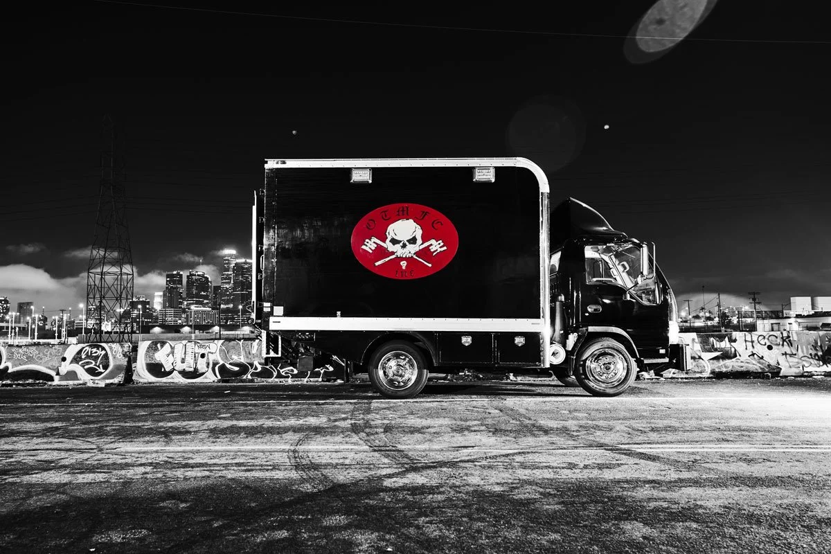 A black delivery truck with a logo featuring a skull, crossed wrenches, and the text 'CUT HUF TUC' in a red circle, parked on an urban street at night with a city skyline and graffiti-covered wall in the background.