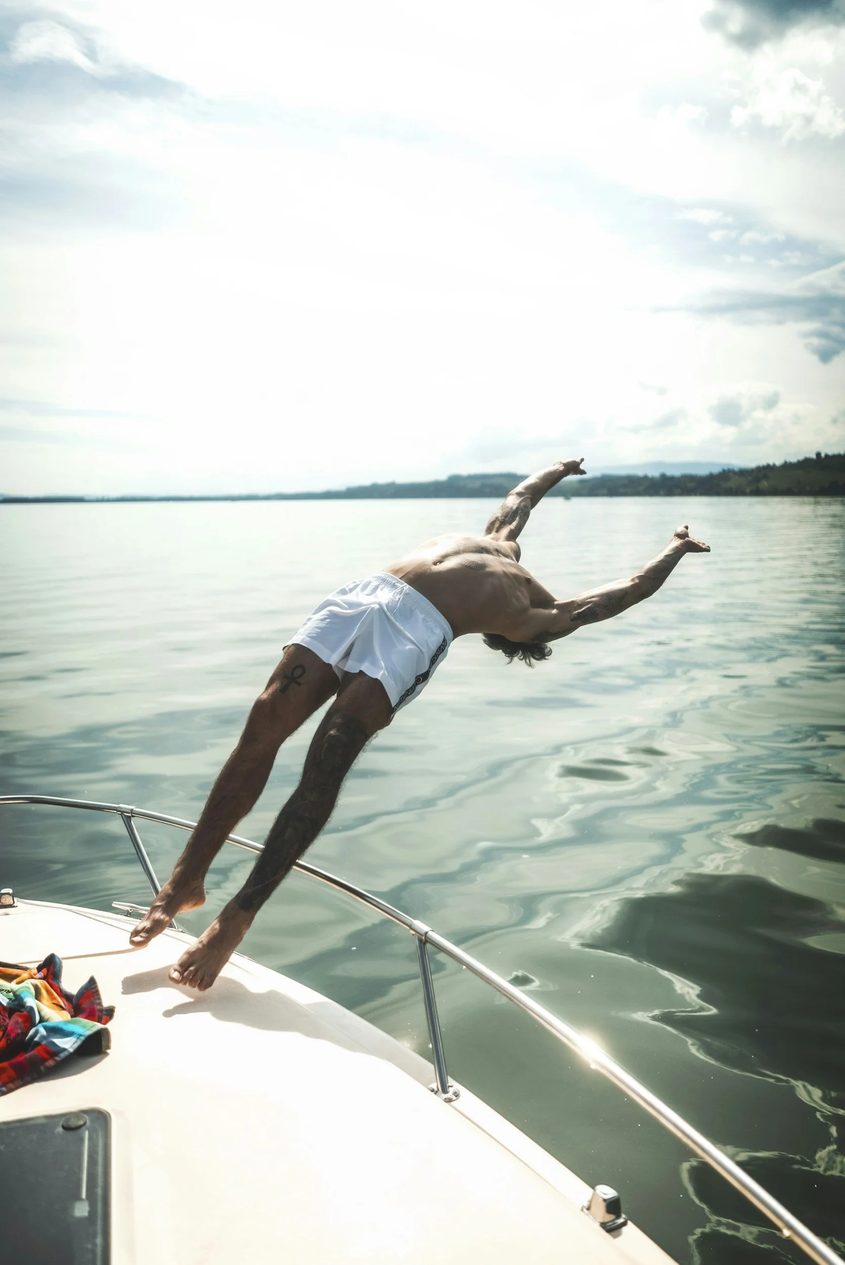 A person diving headfirst off a boat into a calm lake or river, with a partly cloudy sky overhead and distant shorelines.