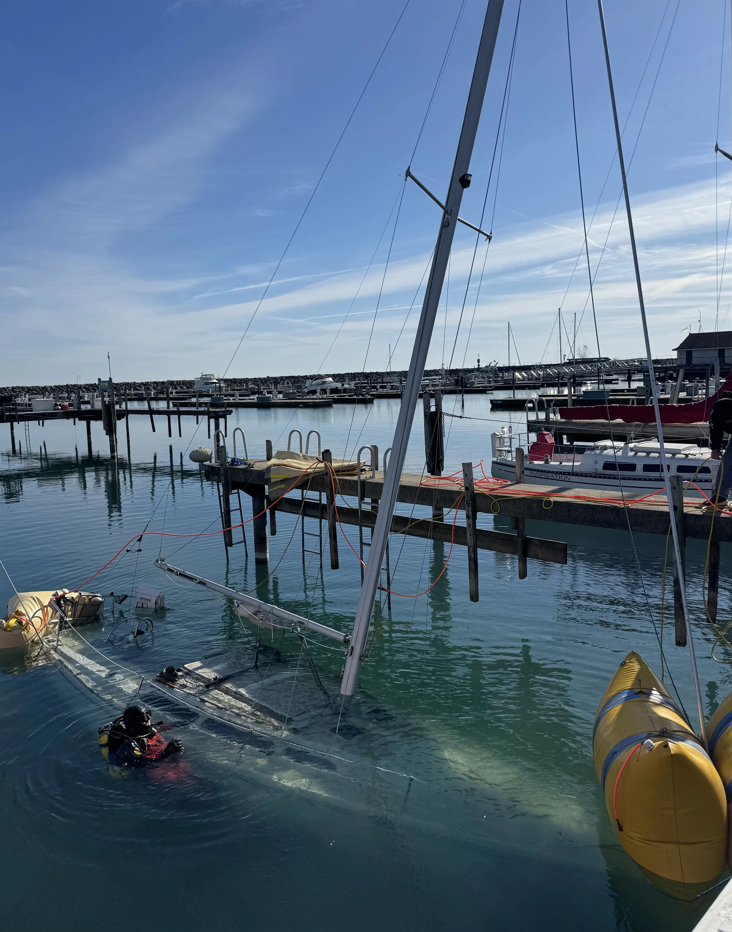 A sailboat has capsized at a marina, with a person in wetsuit in the water near the wrecked vessel, and the mast is partially submerged.