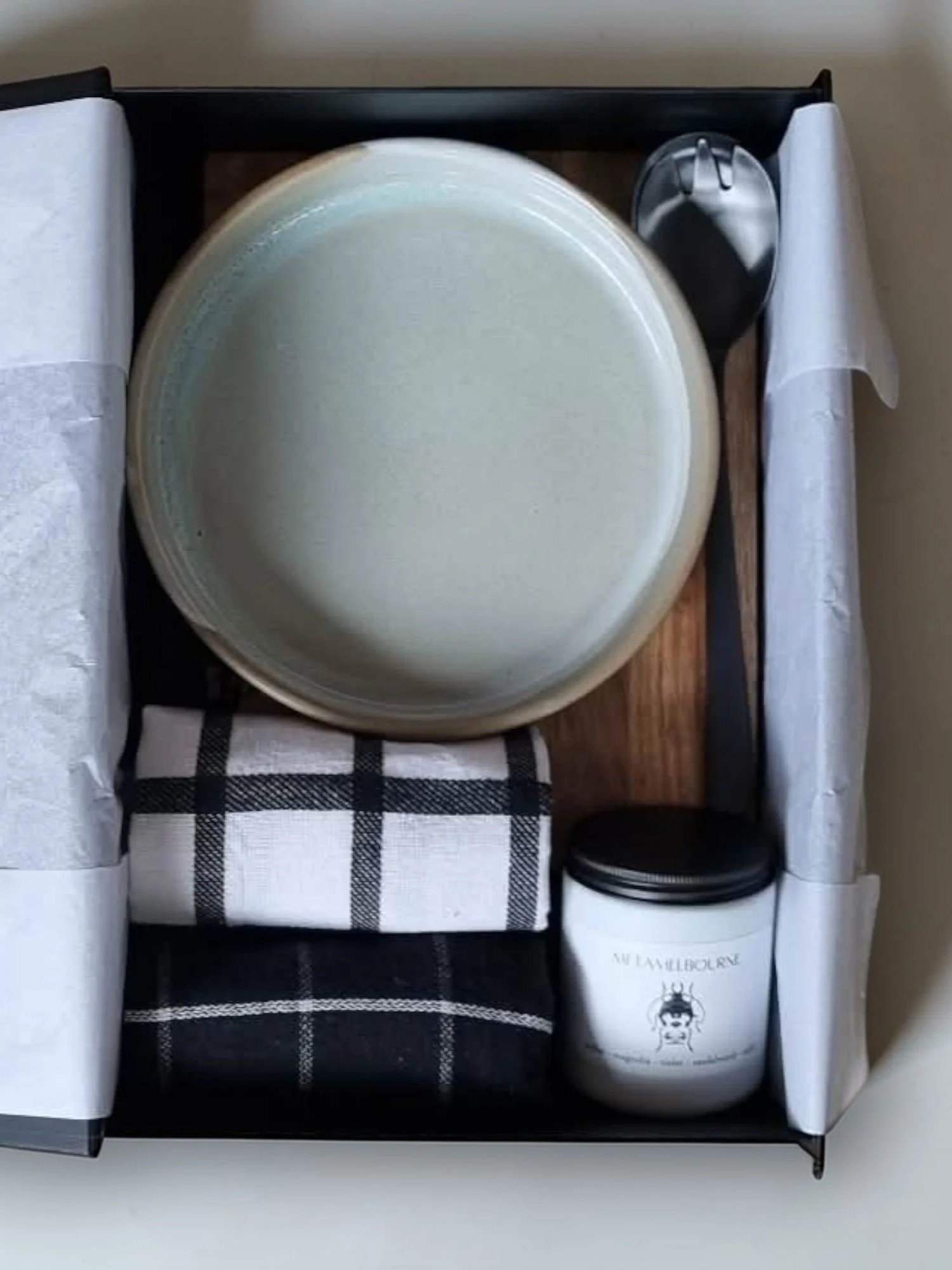 A gift box with white tissue paper, a large round white candle, a smaller white candle with a black illustration and text, a black and white checkered cloth, a black silk ribbon, and a small clear container of cream or lotion.