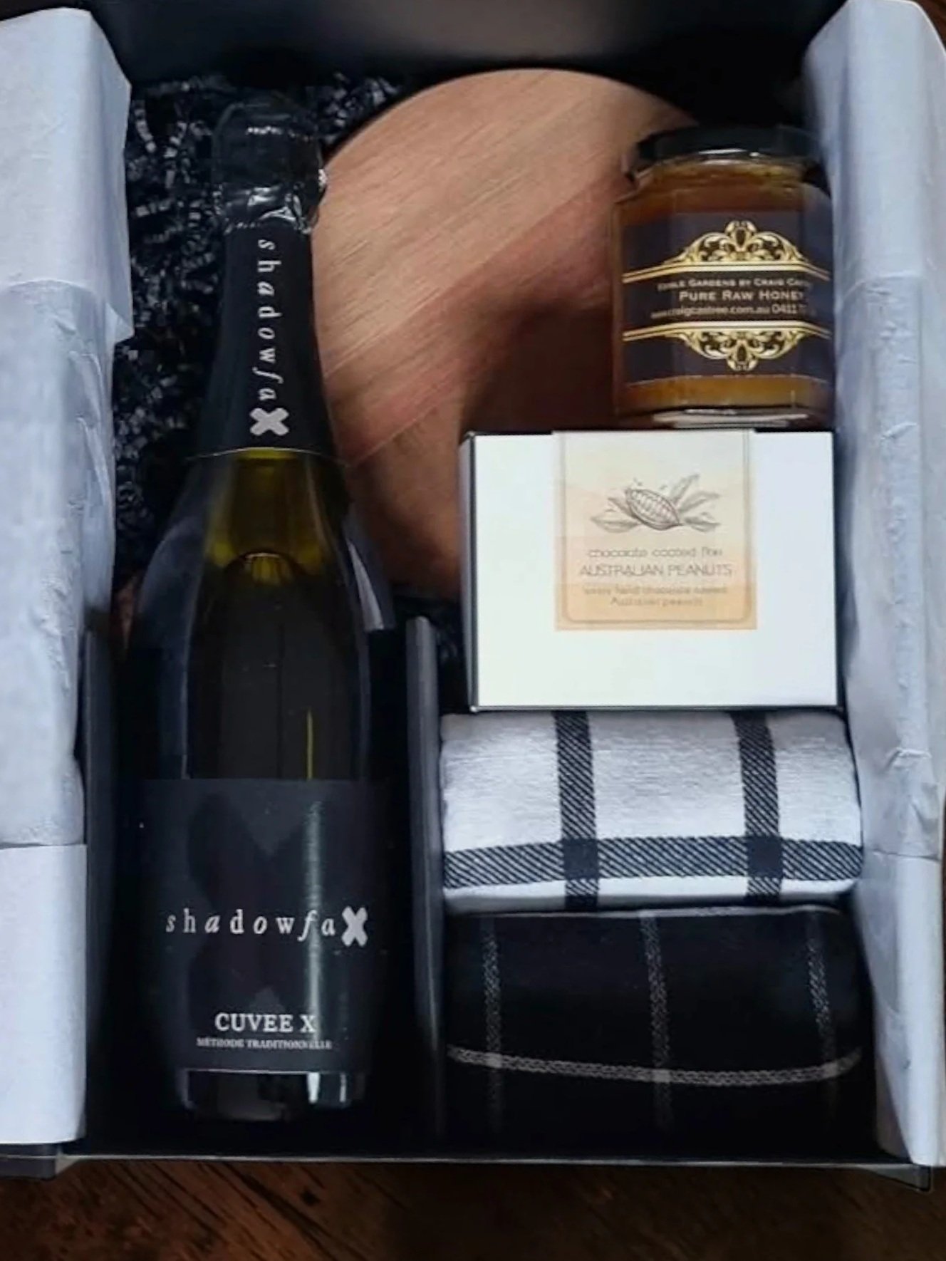A gift box containing a bottle of shadowfax Cuvee X wine, a jar of pure raw honey, a box of Australian peanuts, a wooden spoon, and a plaid cloth napkin.