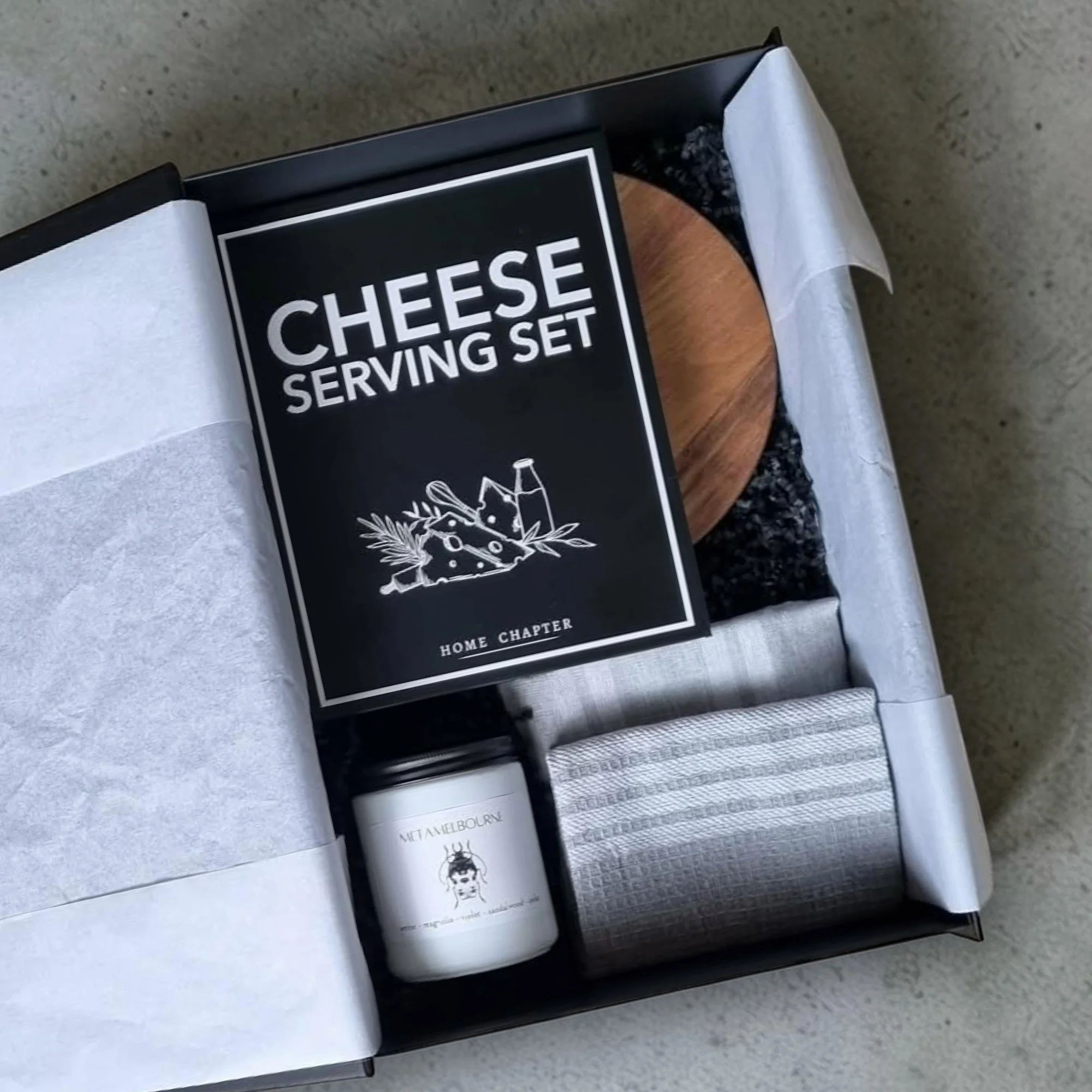 Set of cheese serving tools in black box with black and white label featuring a drawing of a woman's face, placed on a gray surface.