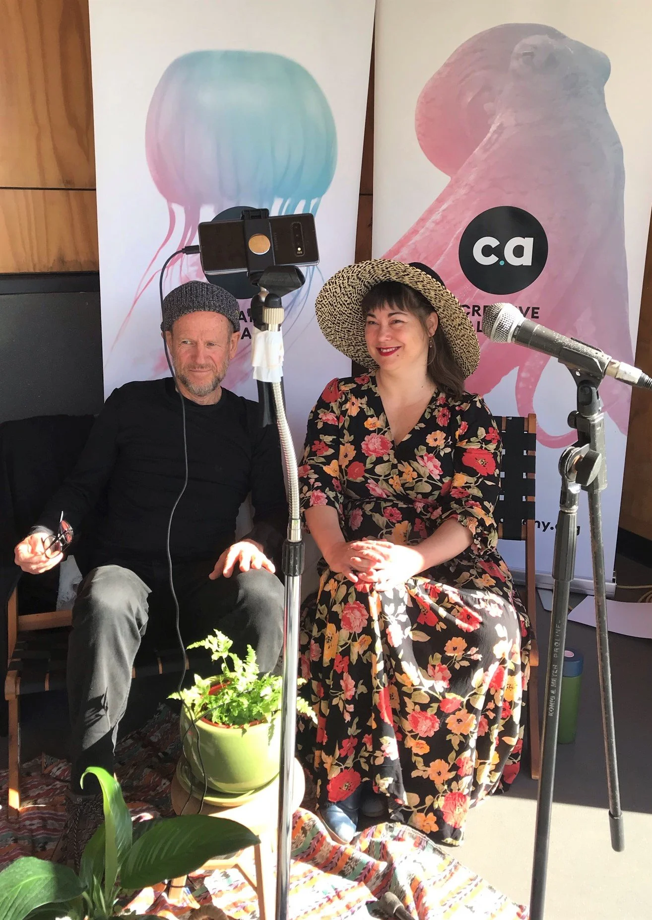 Two people seated indoors, smiling for a photo, with a video recording setup in front of them. Behind them are two vertical banners with abstract art and the letters 'ca' in black circles. The woman on the right is wearing a large straw hat and a flo