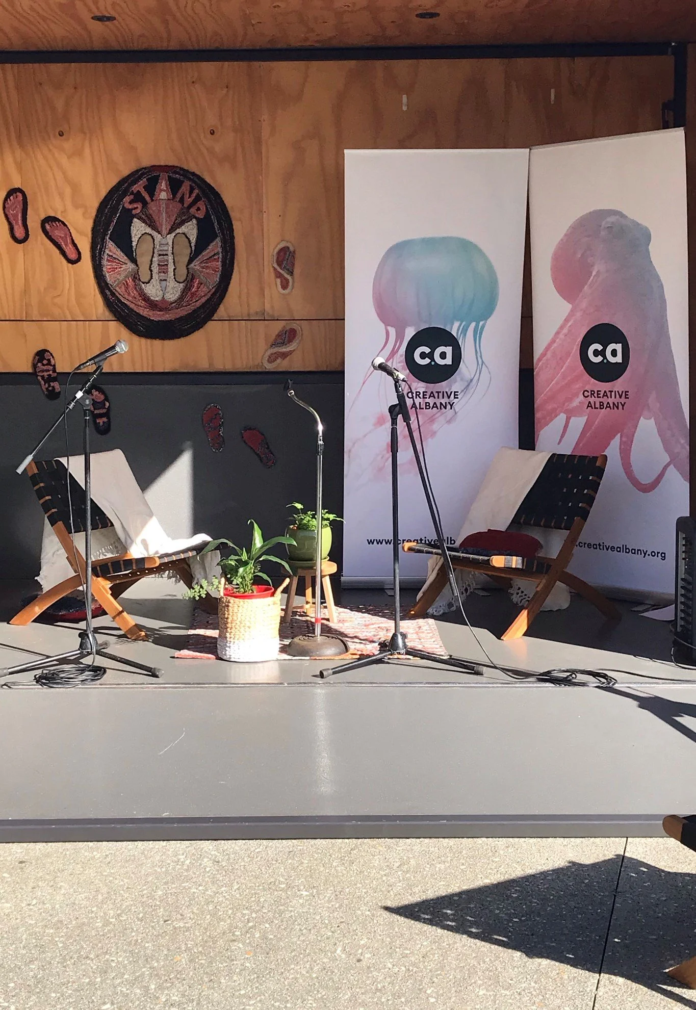 A stage set up for a talk or interview with two chairs, two microphones, and potted plants, with banners reading 'Creative Albany' and a wall decoration featuring a 'Stop' sign with footprints.