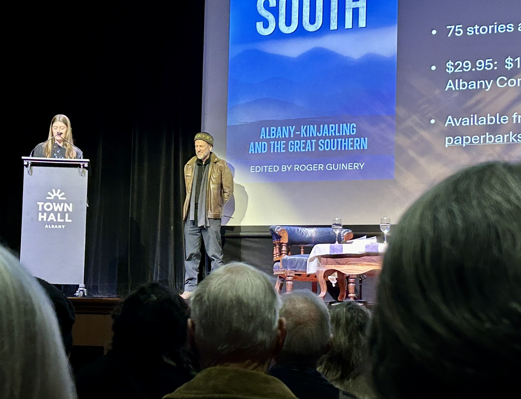 A woman at a podium with the words 'Town Hall Albany' and a man standing beside her on stage. Behind them is a large screen displaying a presentation slide with the title 'South' and details about a book titled 'Albany-Kinjarling and the Great Southe