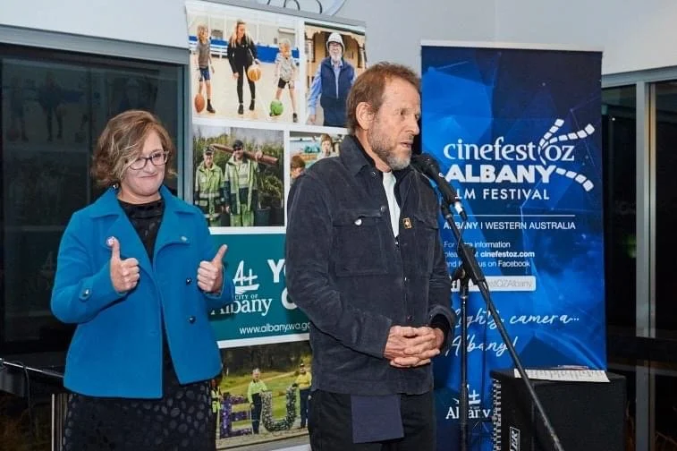 A man speaking at a microphone with a woman standing beside him giving a thumbs up, at an outdoor event for the Albany Film Festival in Western Australia. There are banners and posters in the background.