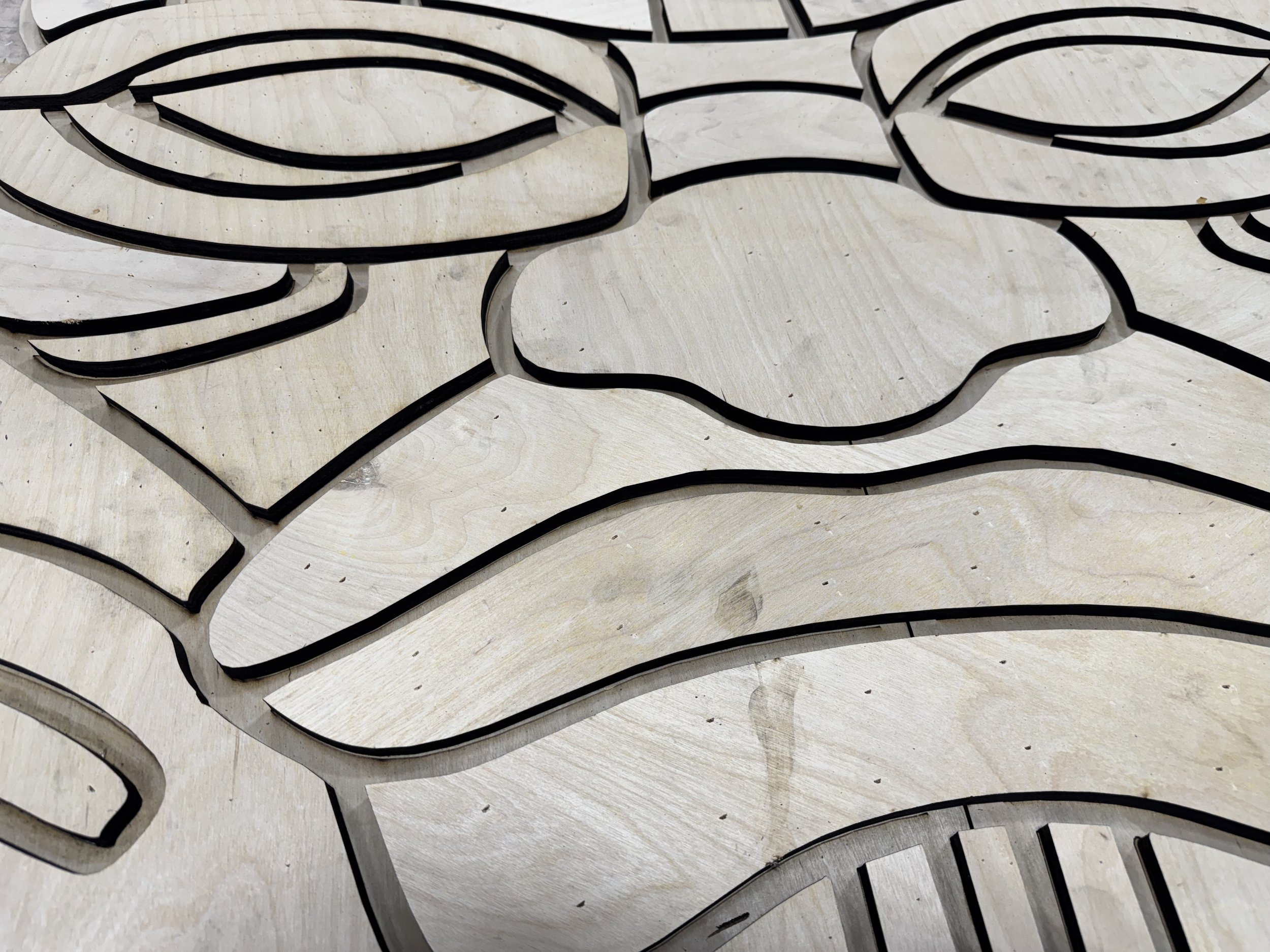 Laser-cut plywood parts with dark scorched edges, nested tightly together in an organic interlocking pattern on the shop floor.