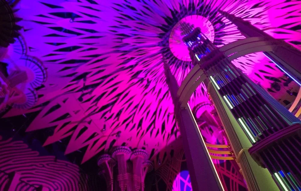 Looking up at the Atlas 9 ceiling mandala, laser-cut petals radiating outward under magenta light, with the central spire and an arched catwalk crossing the frame.