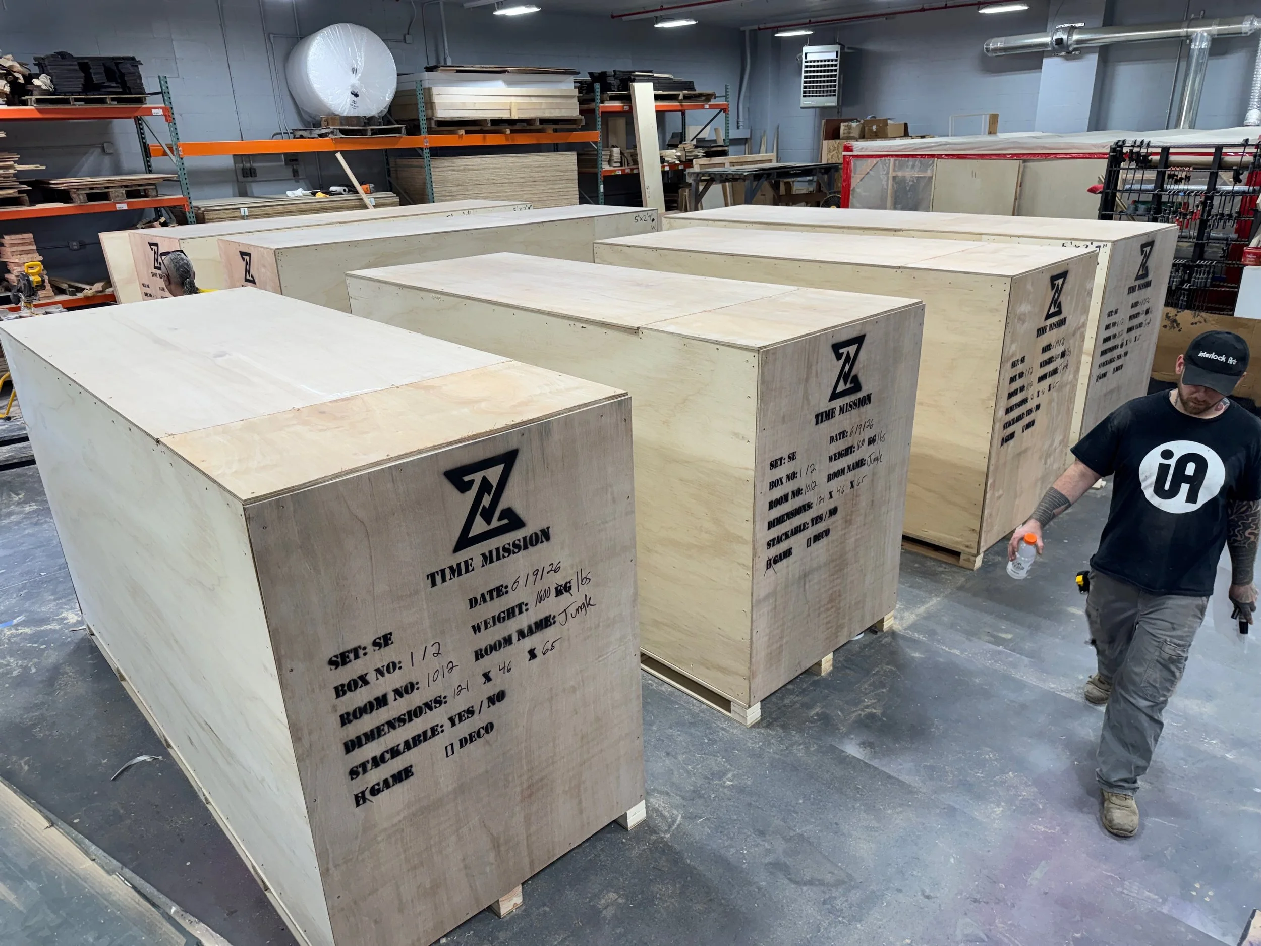 Six plywood shipping crates stenciled with the Time Mission logo and labeled by set, box number, room name, weight, and dimensions, staged for freight on the shop floor with a crew member walking past.