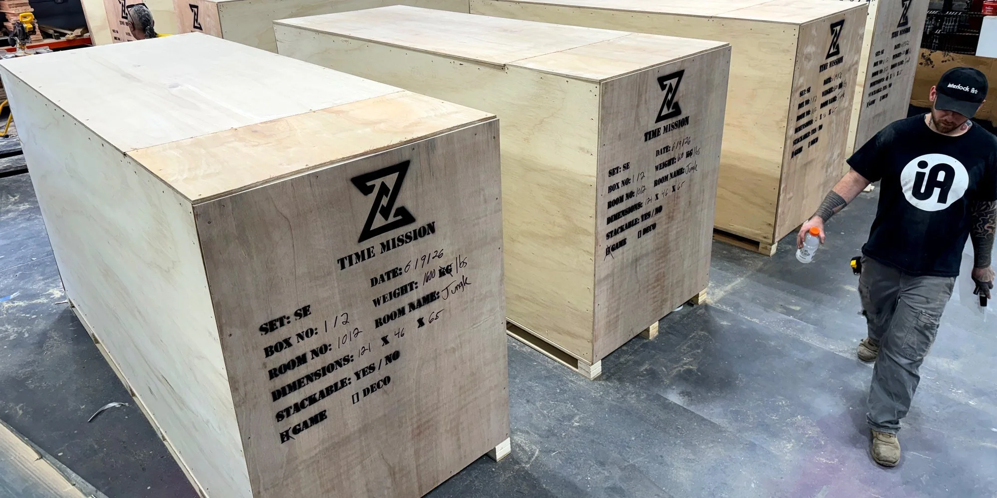 Six plywood shipping crates stenciled with the Time Mission logo and labeled by set, box number, room name, weight, and dimensions, staged for freight on the shop floor with a crew member walking past.