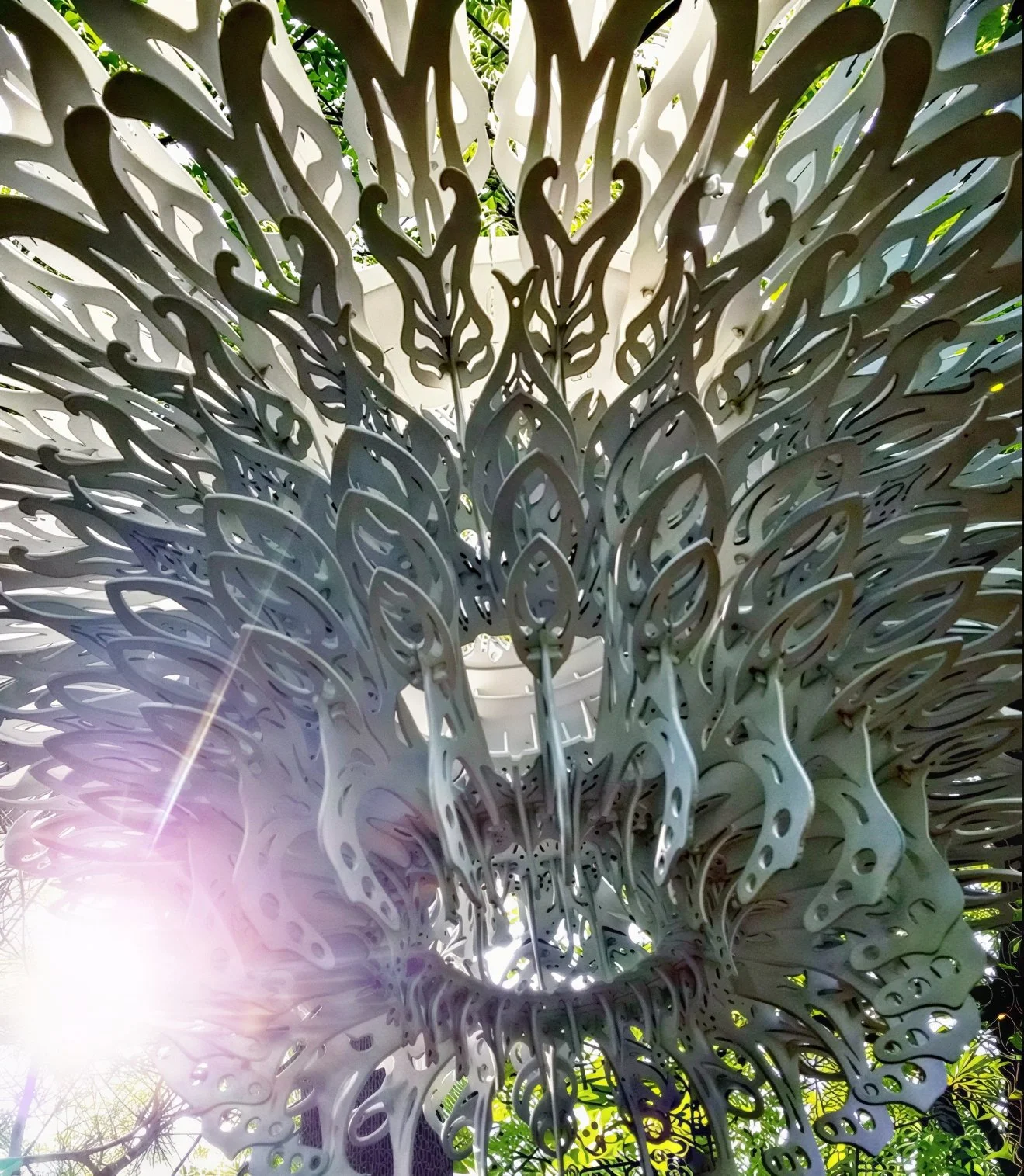 Looking up through the Terra Luna chandelier in daylight, sunlight streaming between the white laser-cut layers with green leaves visible beyond.