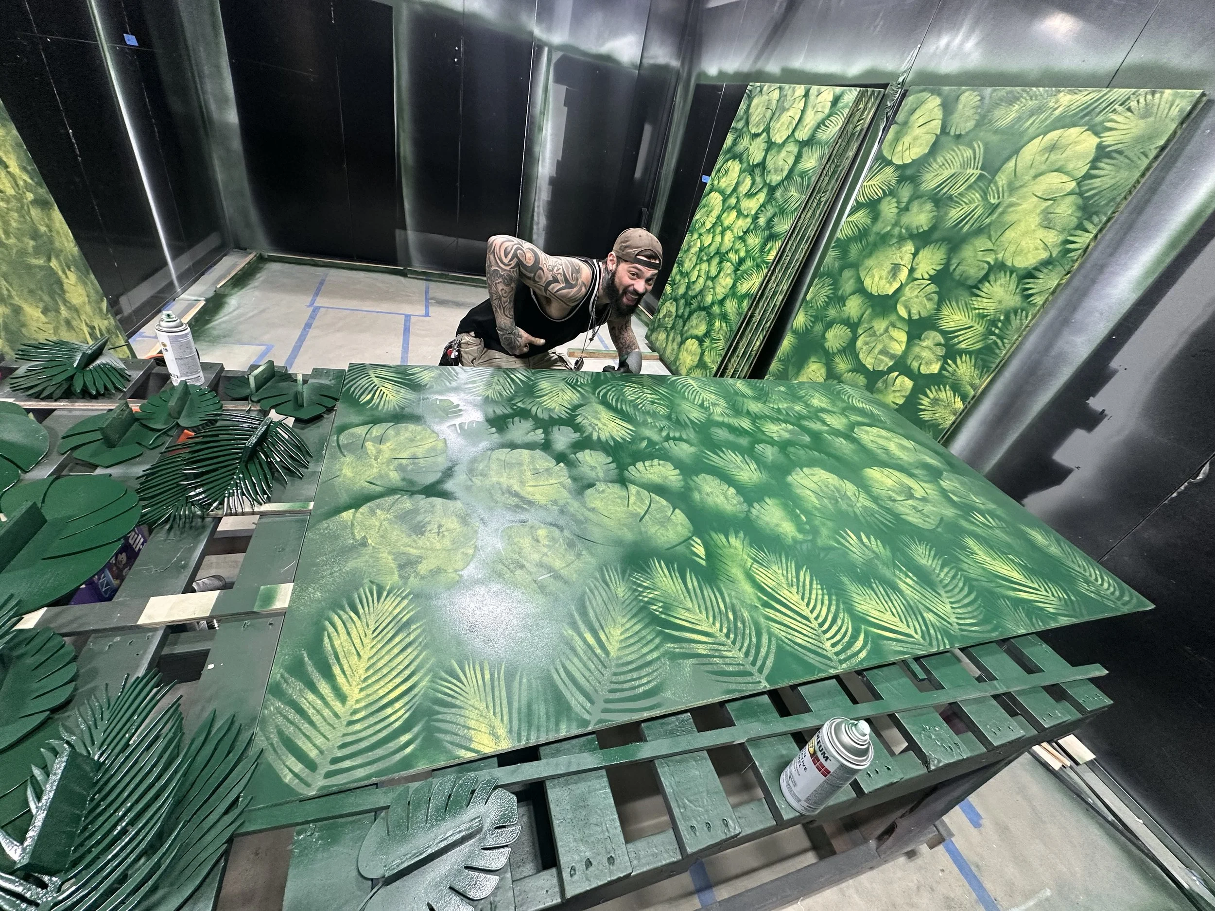 A finisher spray-painting laser-cut fern and palm panels in the studio's paint booth, with finished green panels stacked behind him.