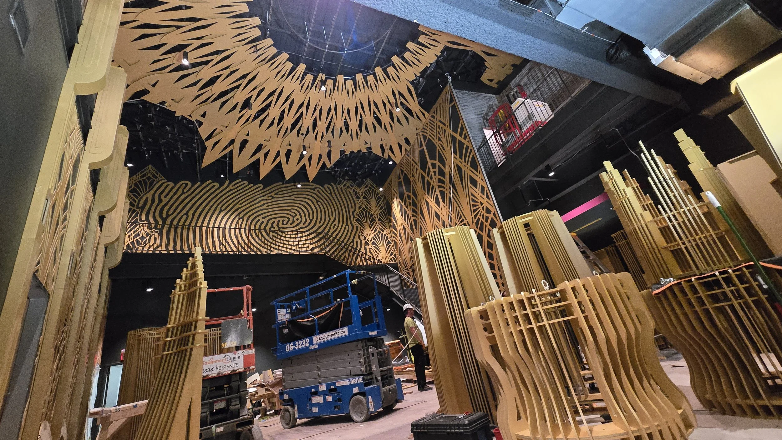 The Atlas 9 ceiling mandala mid-installation in raw plywood, with a scissor lift on the floor below and stacks of cut spire components waiting to be assembled.