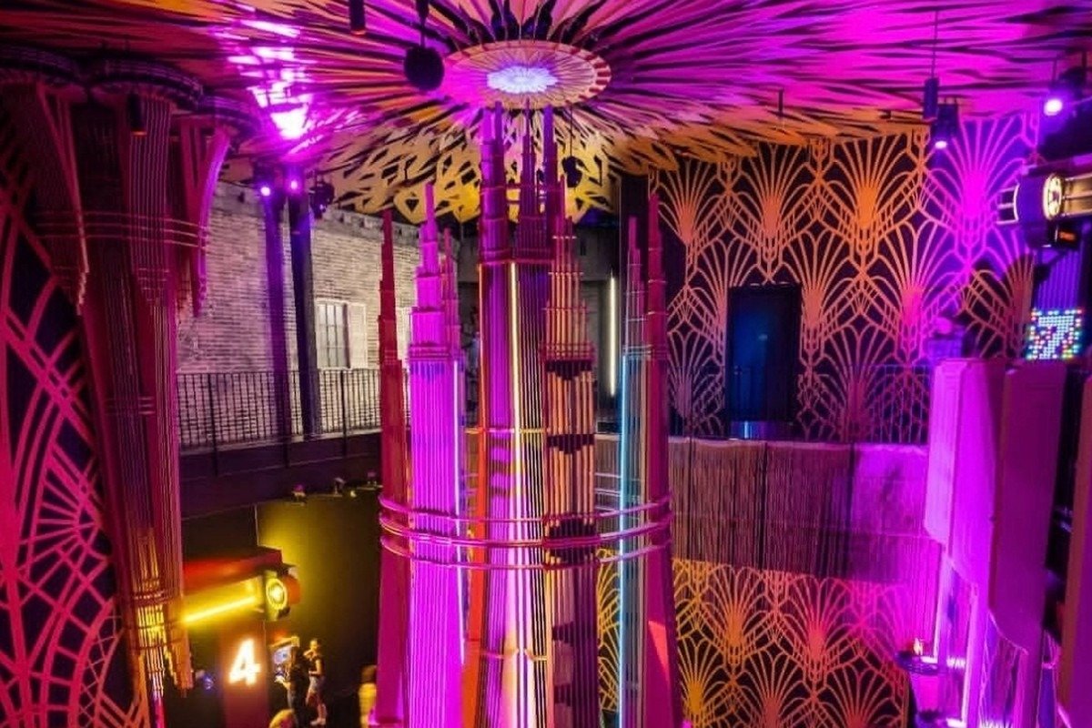 Magenta-lit jazz club interior with a laser-cut ceiling mandala radiating overhead and a tall central spire rising from the floor, guests below for scale.