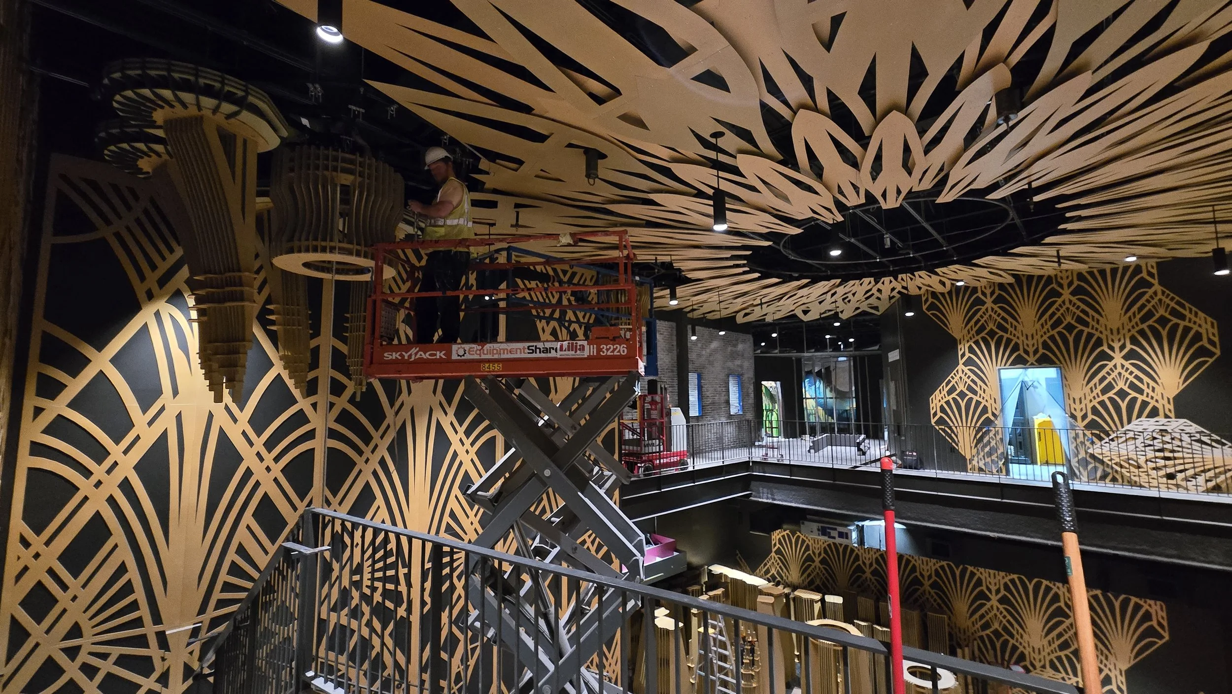 A crew member on a scissor lift installing the unfinished Atlas 9 ceiling mandala, laser-cut plywood petals radiating overhead above the deco wall panels below.