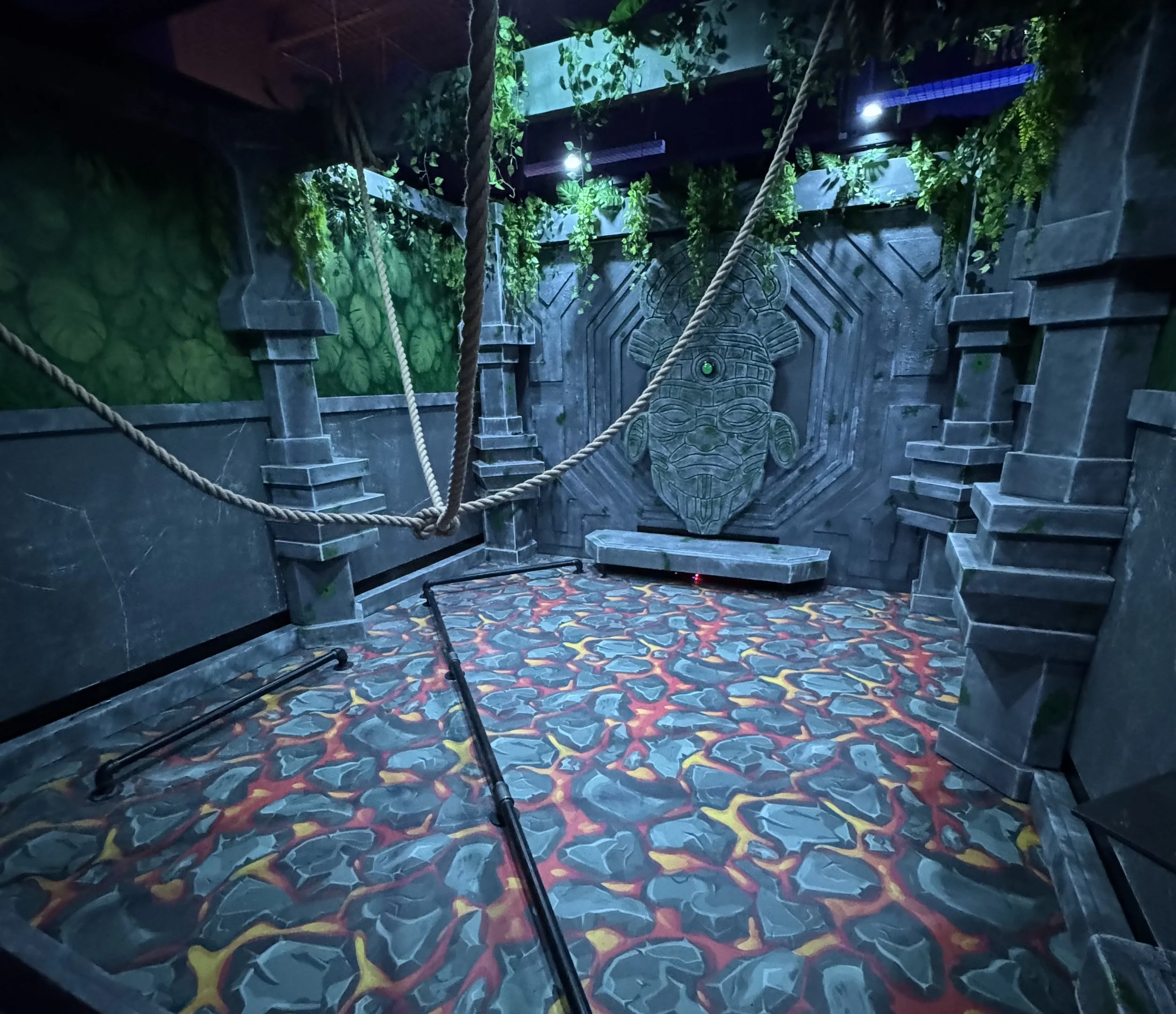 Immersive escape-room set built as a stone temple chamber, with hanging ropes, a carved mask relief, and a hand-painted lava-rock floor.