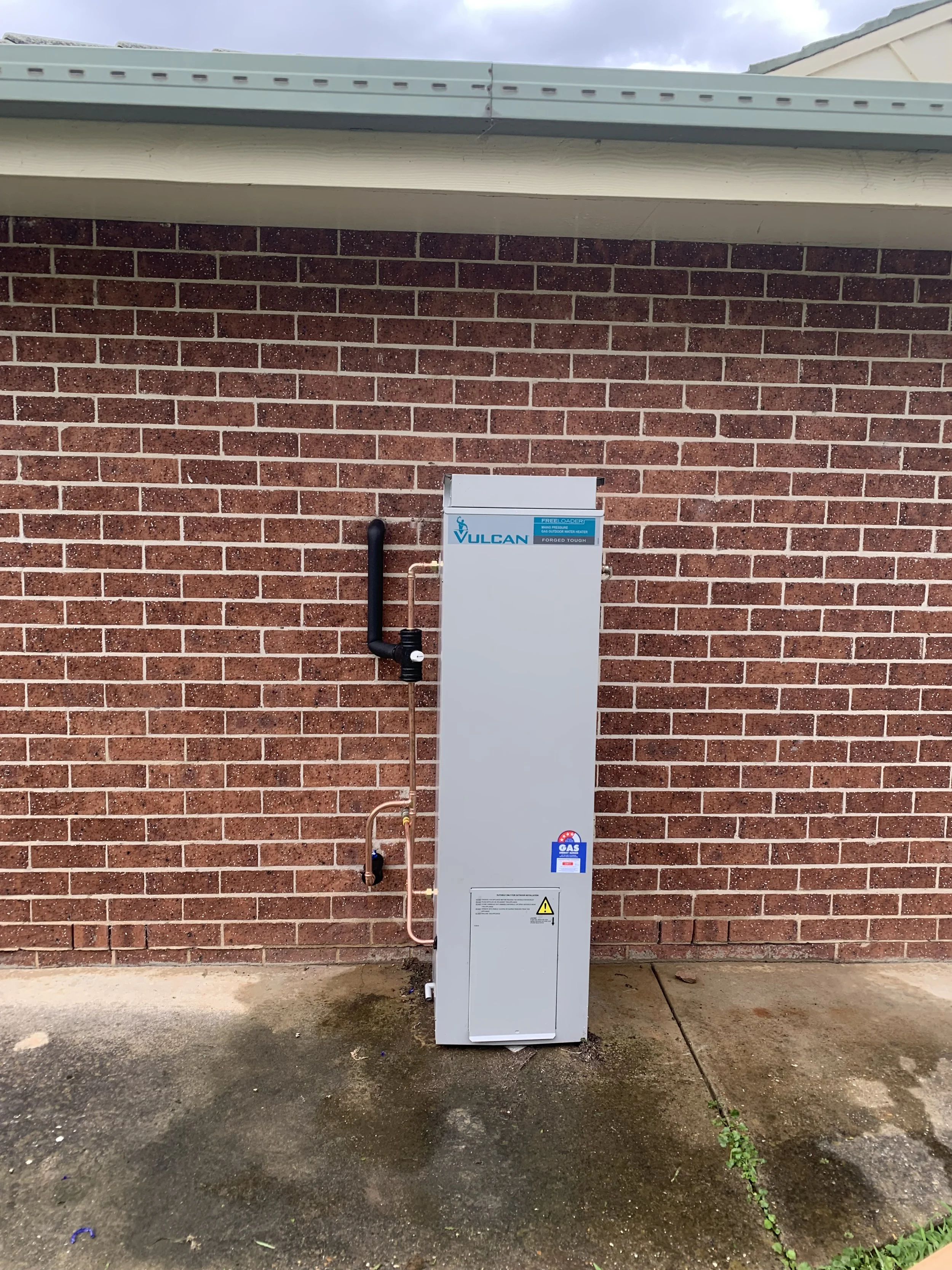 External view of a Vulcan LPG gas tank system installed against a red brick wall, with pipes and warning labels.