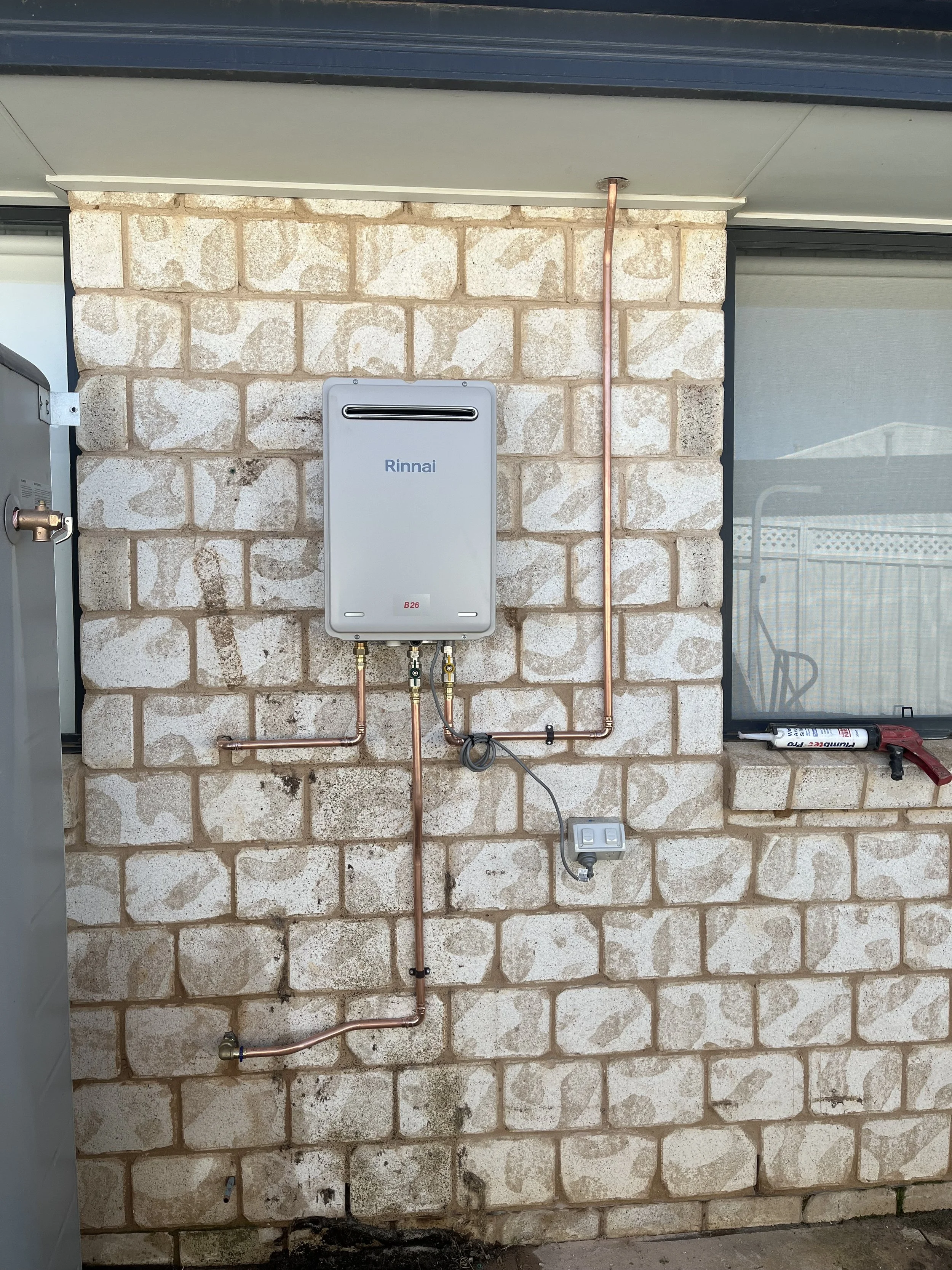 Outdoor Rinnai tankless water heater mounted on brick wall, with copper pipes connected, a small gray control box, and a window with a garden tool and a tube nearby.