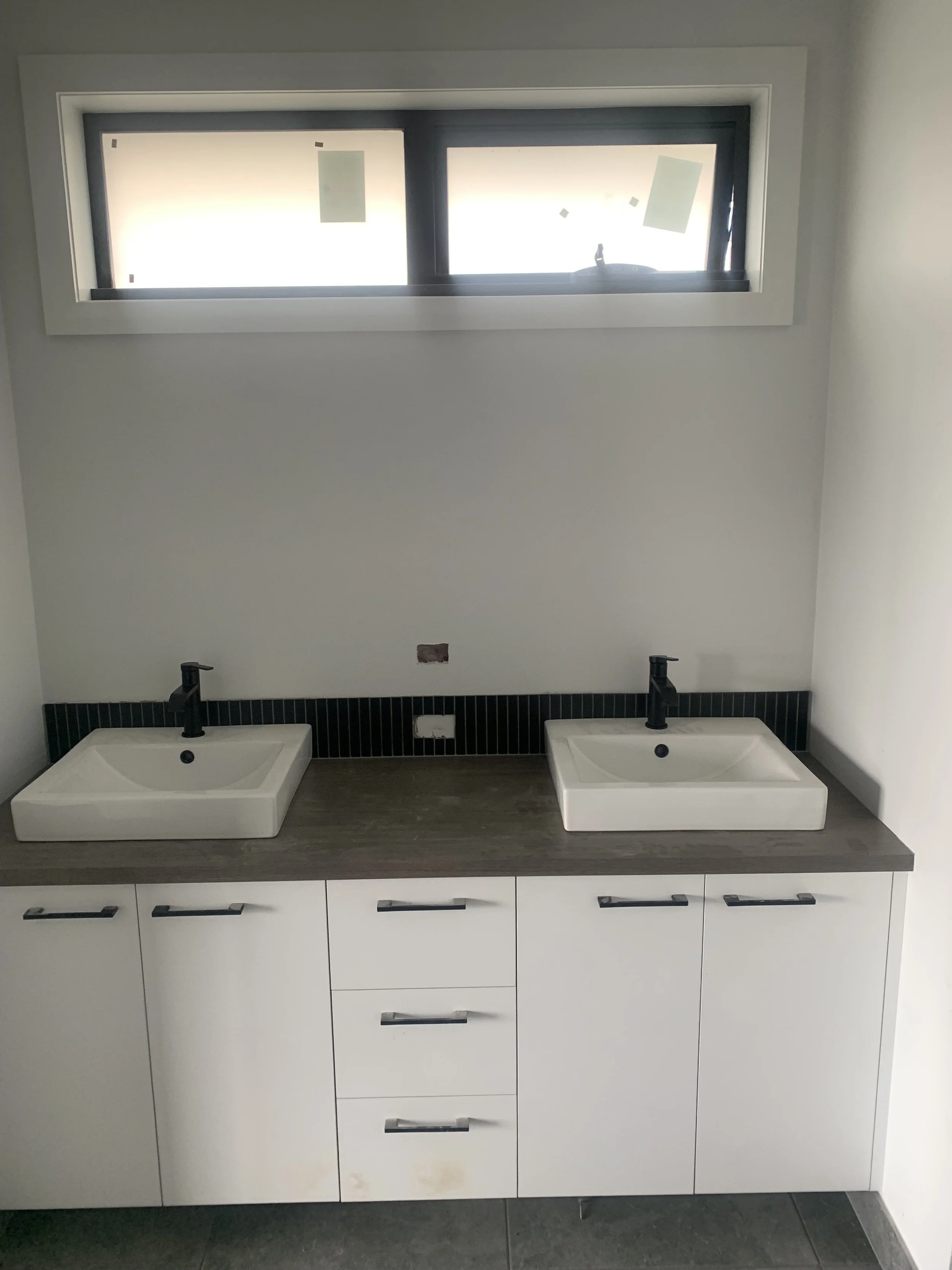 Bathroom with two square sinks on a wooden countertop, black faucets, white cabinets, window above, and a black tiled backsplash.