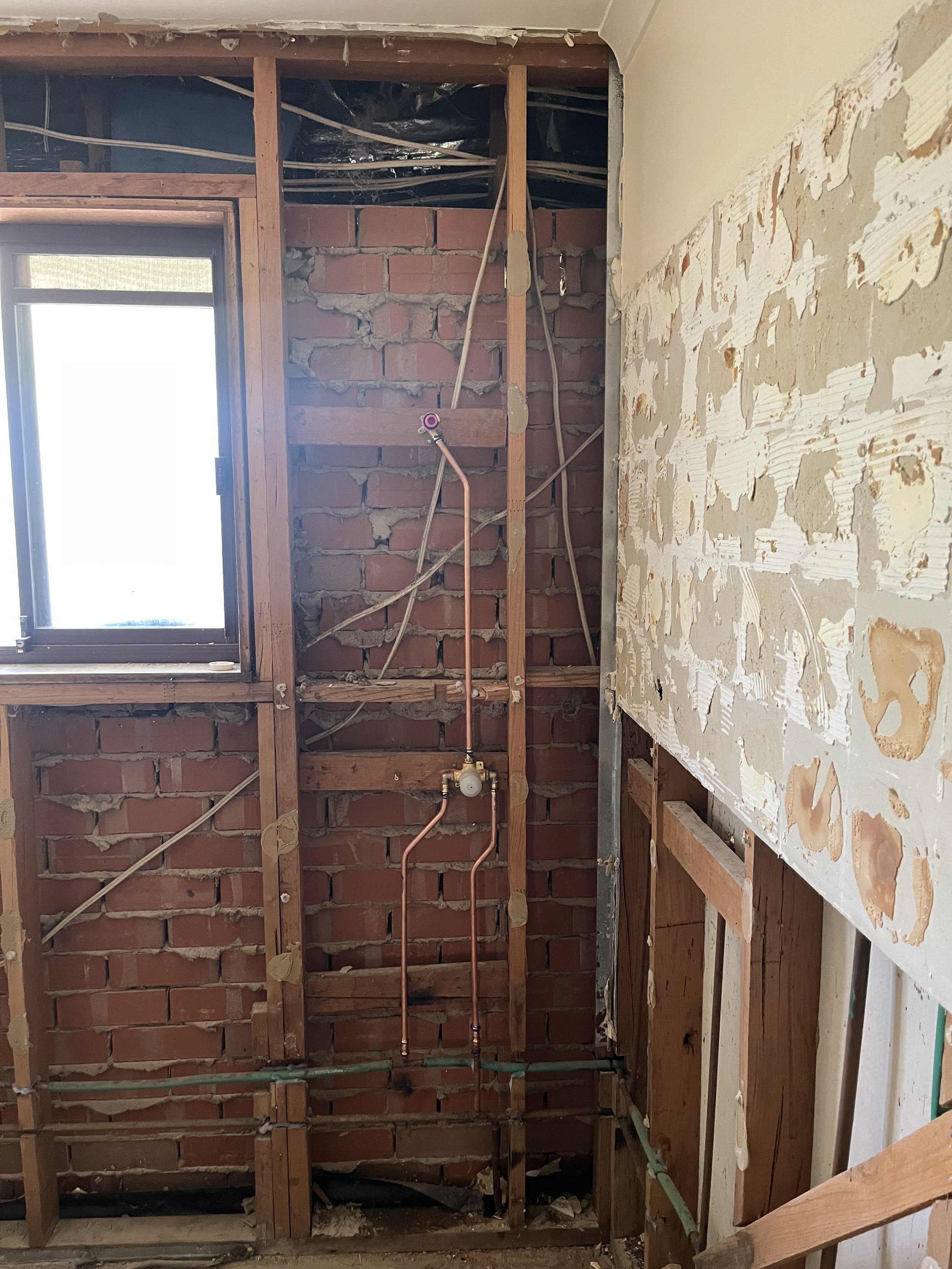Interior view of a room under renovation with exposed brick wall, electrical wires, plumbing pipes, and a window.