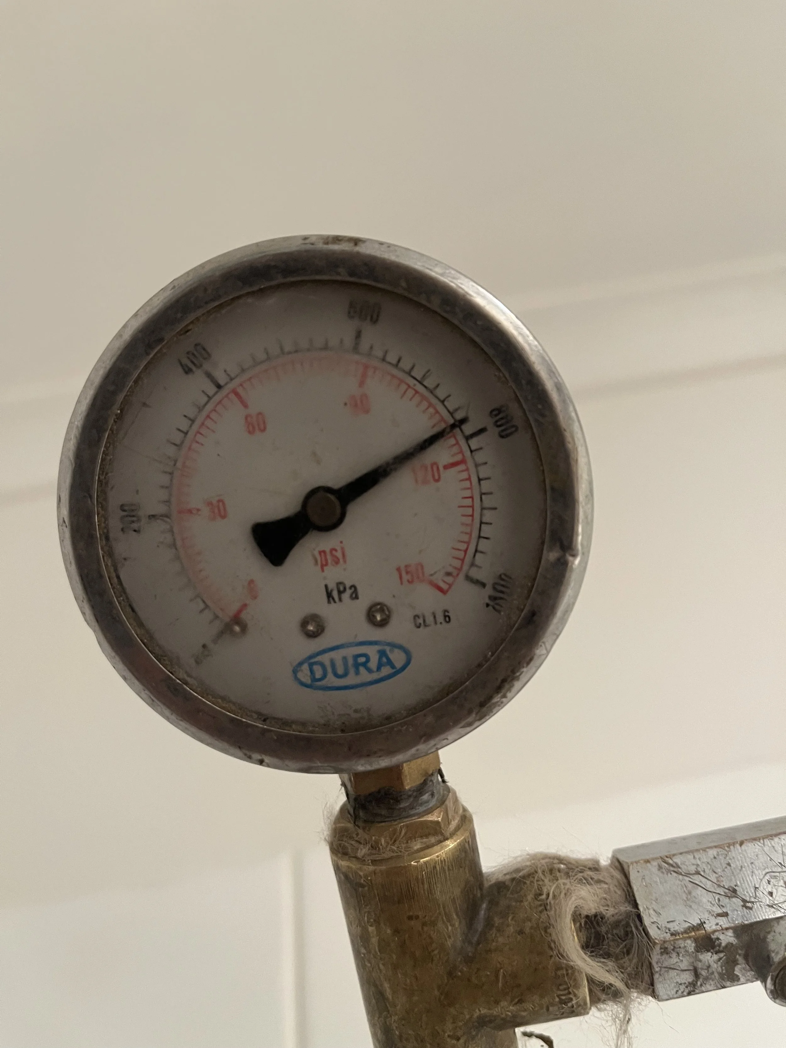 A pressure gauge showing a pressure of approximately 150 psi, manufactured by DURA, mounted on a metallic pipe with some worn material around the connection.
