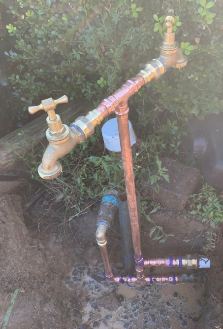 A backyard water faucet organization setup with copper and brass pipes, valves, and a garden hose connection amid grass and rocks.