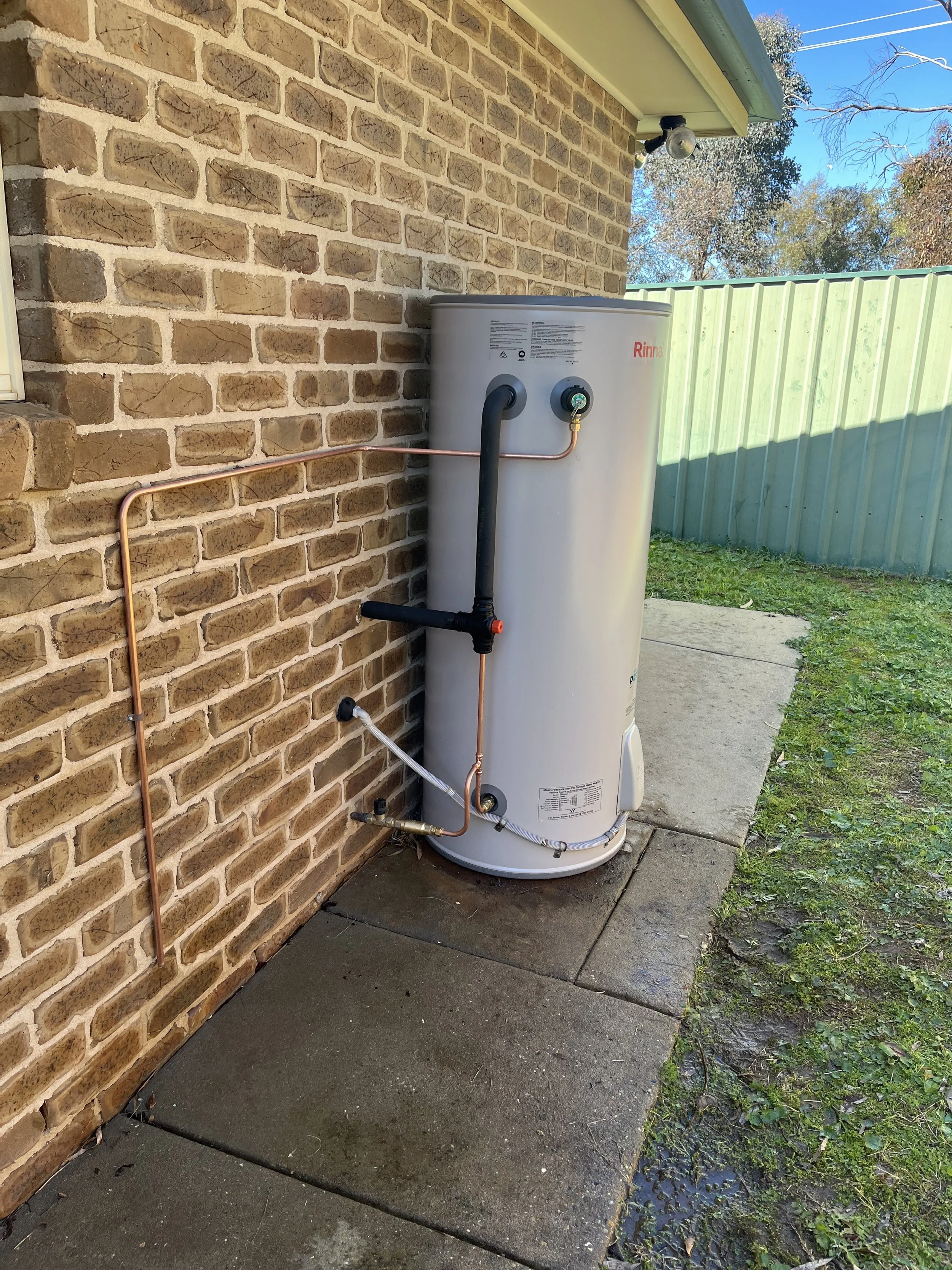 Outdoor water heater installed next to a brick wall, with copper and black piping connected to it, situated on concrete panels with grass nearby, and a green fence in the background.